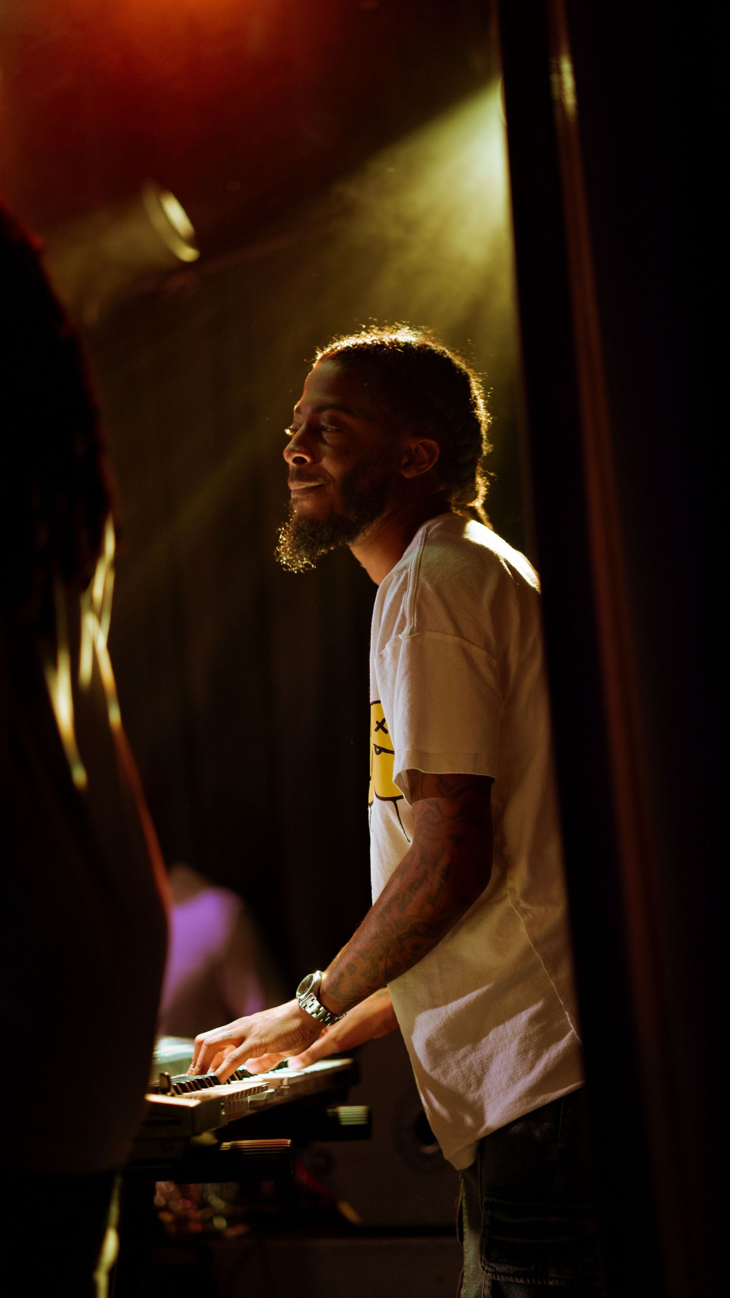 Smiling male DJ in white t-shirt works equipment under warm stage lighting.