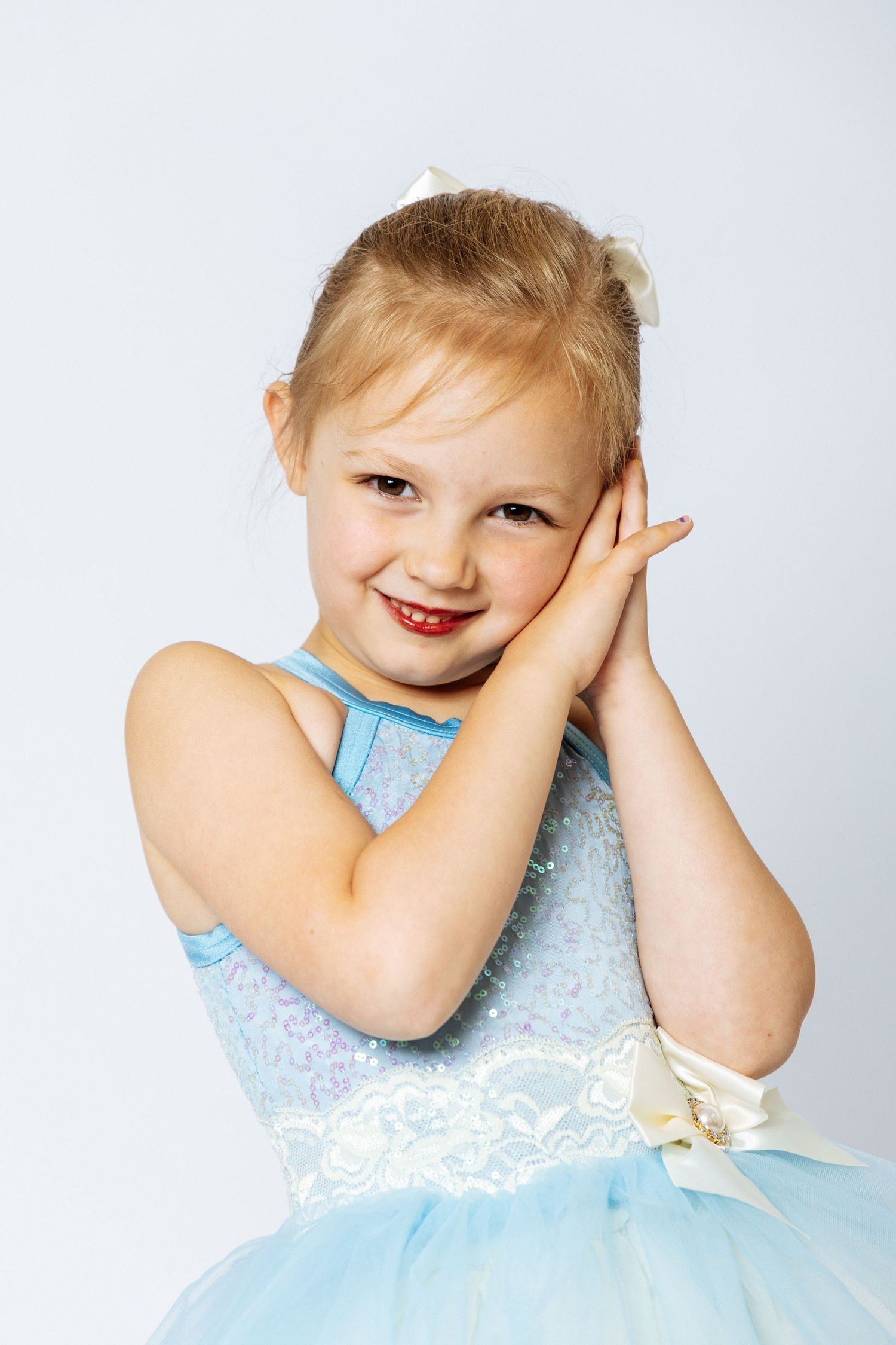 A smiling young girl in a blue tutu dress with a bow, hands pressed together near her cheek, posing against a white background.