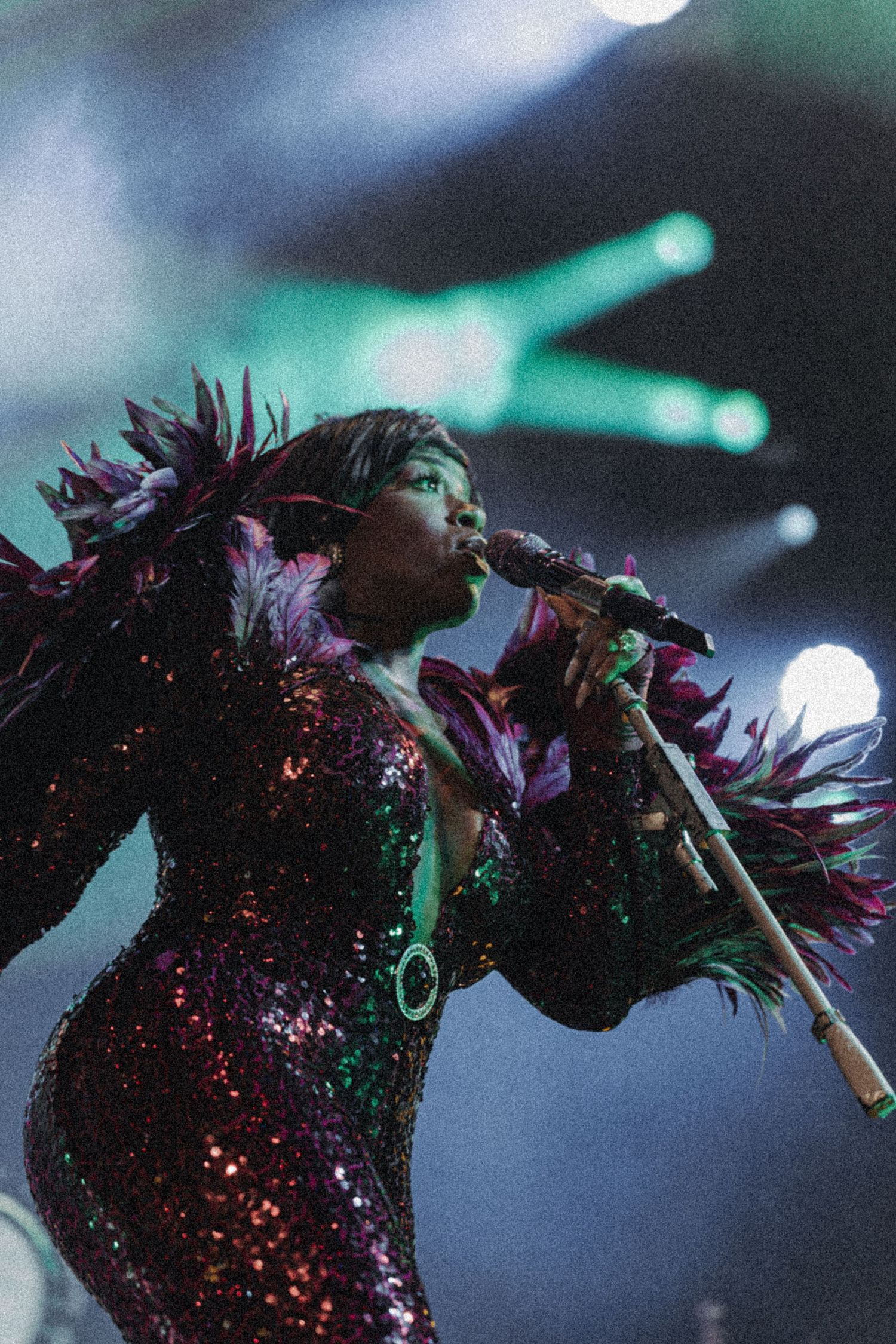 A glittering performer in a sequined outfit with purple feathers sings passionately on a concert stage under dramatic lights.