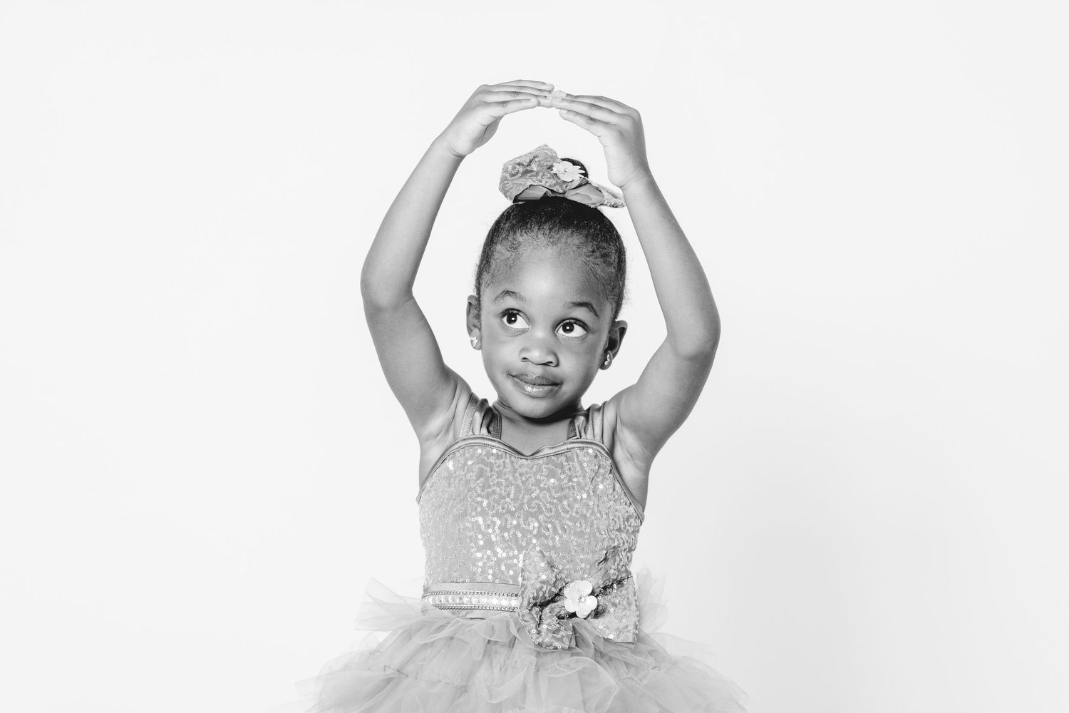 Young Black girl in sparkly tutu dress poses gracefully with arms raised overhead in black and white ballet portrait.