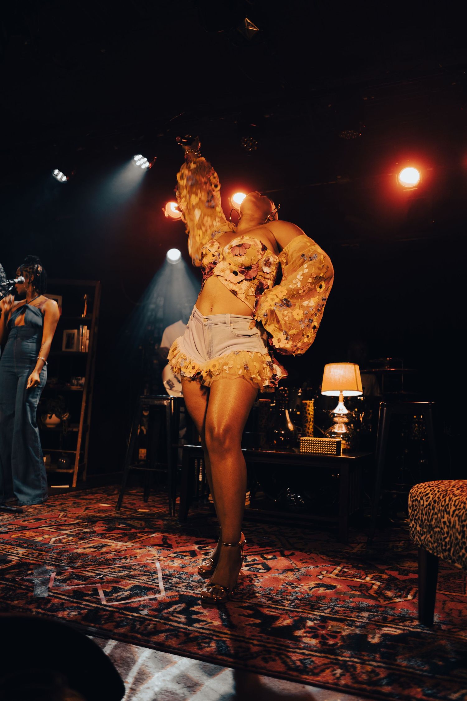 Female performer dancing on stage in gold floral top and denim shorts under dramatic spotlights with vintage decor backdrop.