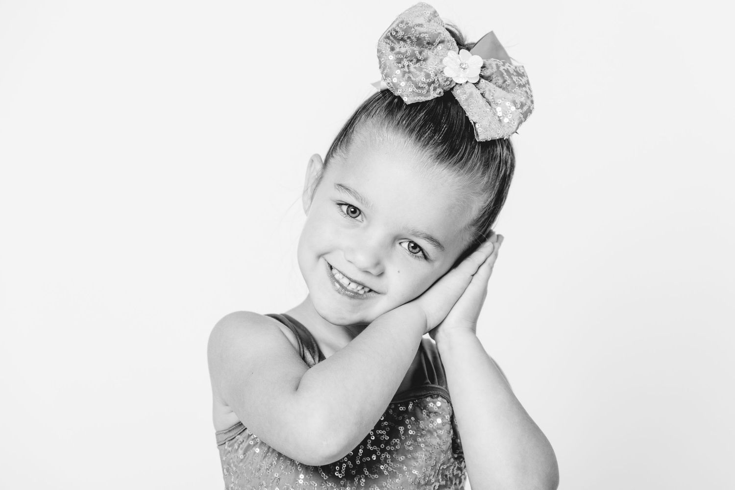 Black and white portrait of a smiling young girl with a bow in her hair, hands on cheeks, wearing a sparkly outfit.