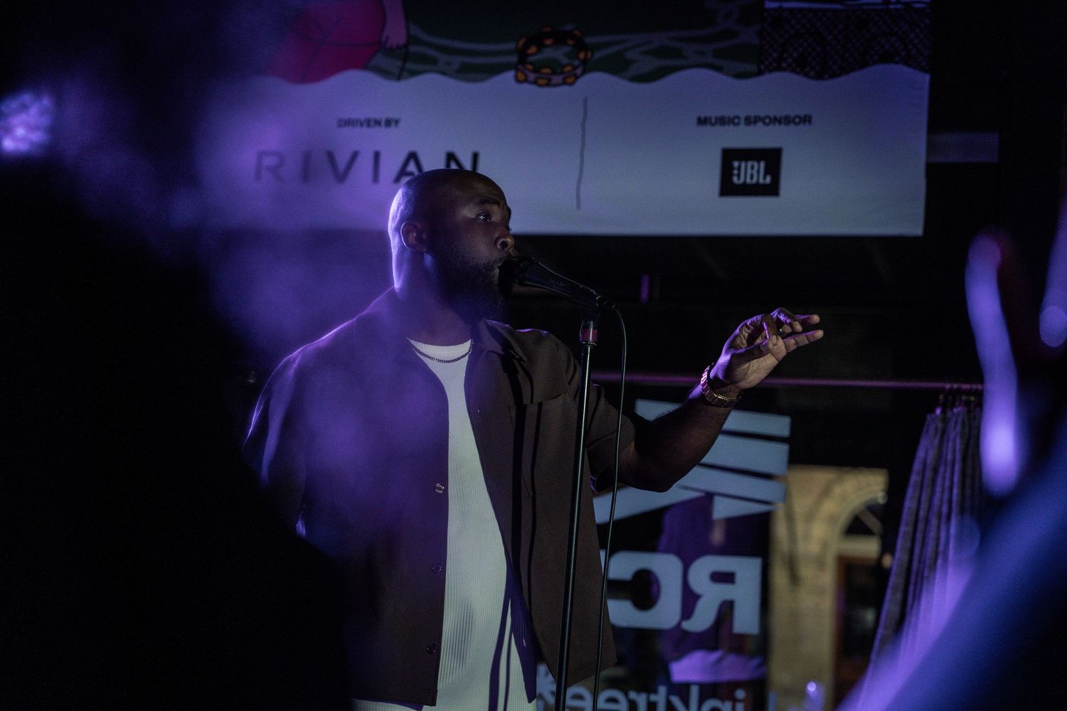 A performer sings into a microphone on a dimly lit stage with Rivian and JBL banners in the background.