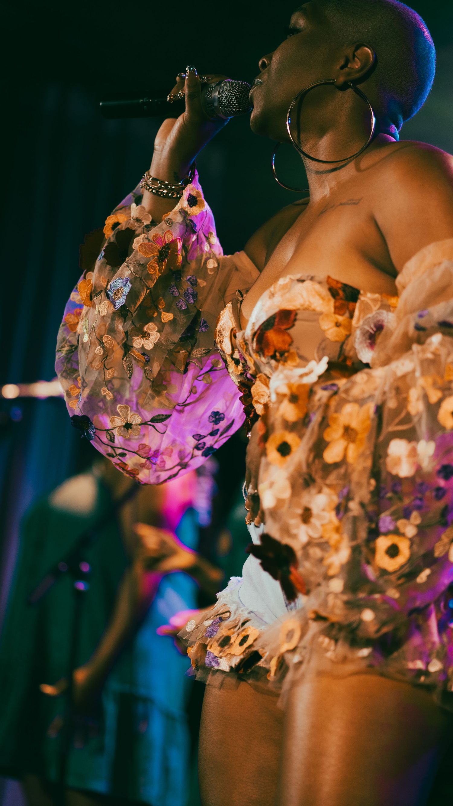 A female singer in a floral sequined outfit performs with a microphone under colorful stage lights.