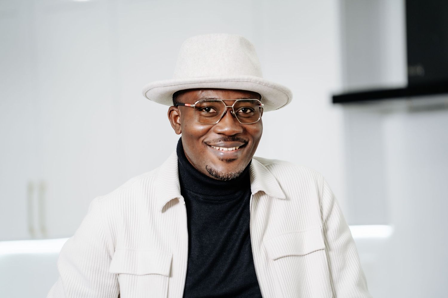 A smiling figure wearing glasses, a white fedora hat and blazer with a black turtleneck in a bright setting.