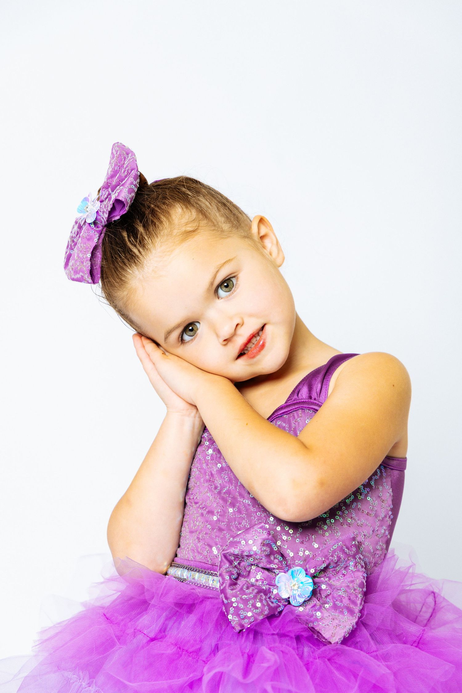 A cute little girl in a purple sequin tutu dress and bow poses with hands on cheeks against a white background.