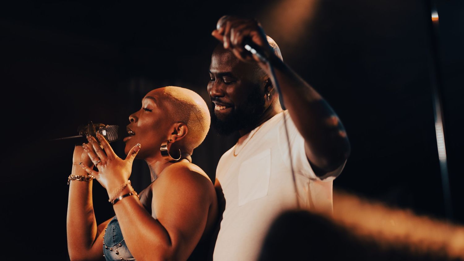 Two singers perform together on a dark stage, a woman and a tall man sharing a microphone during a live concert.