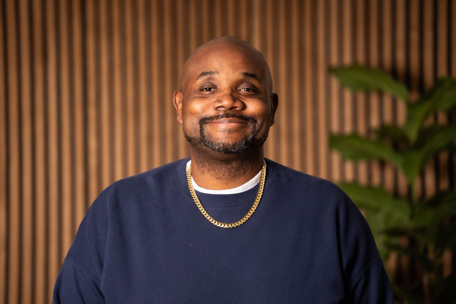 Person wearing a navy blue sweater and gold chain necklace smiles warmly against a wooden slat wall with greenery.