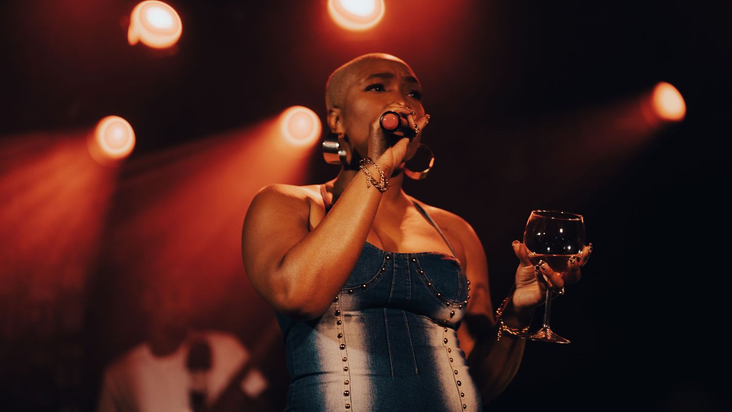 Bald performer in denim outfit holding wine glass on stage under warm red concert lighting.