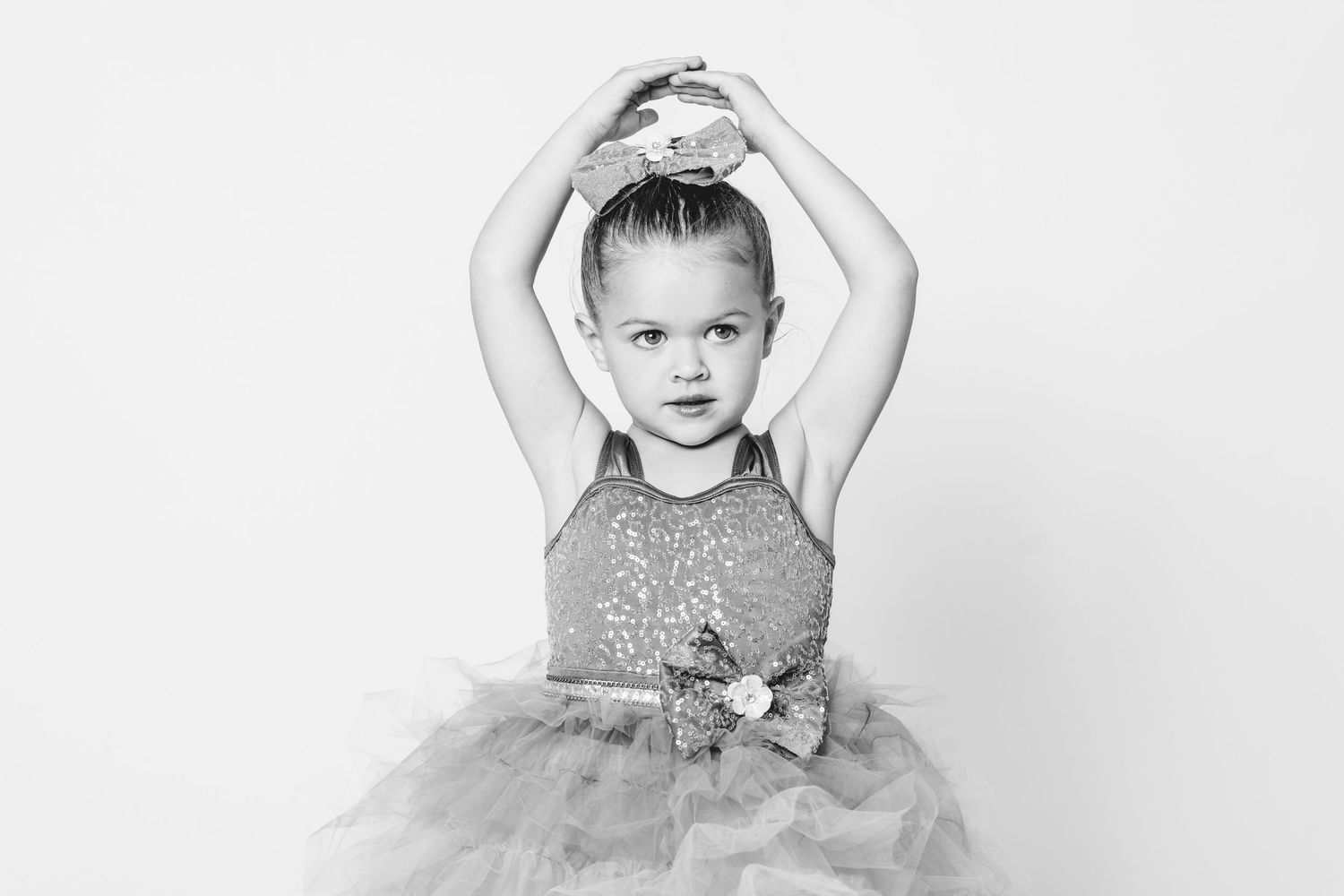 Young girl in sequin dress poses ballet arms overhead in black and white portrait photo.