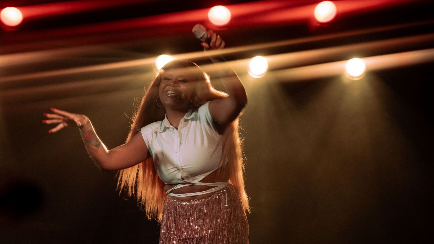 A young female singer performs on stage under warm lights wearing a pink skirt and white top.