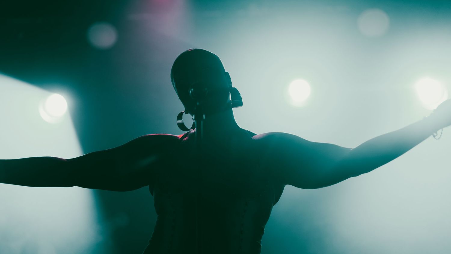 Shirtless male performer on stage with arms outstretched bathed in blue-green spotlight from behind.