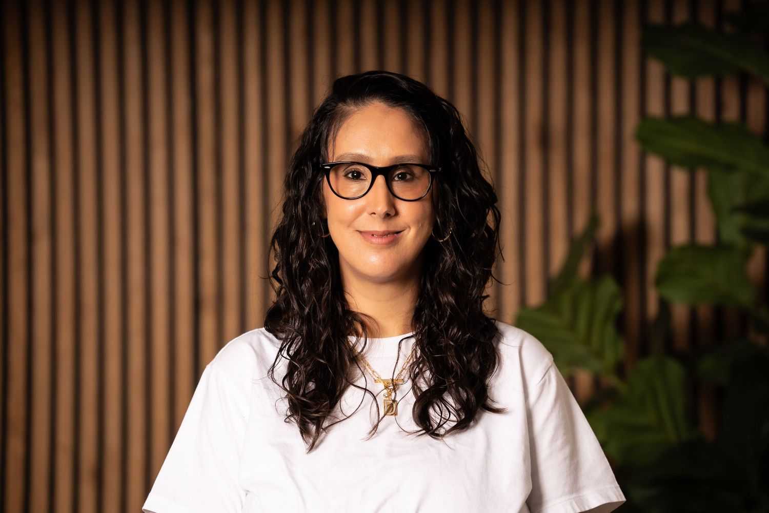 Professional headshot portrait of someone with curly dark hair and glasses wearing a white blouse against a wooden background.