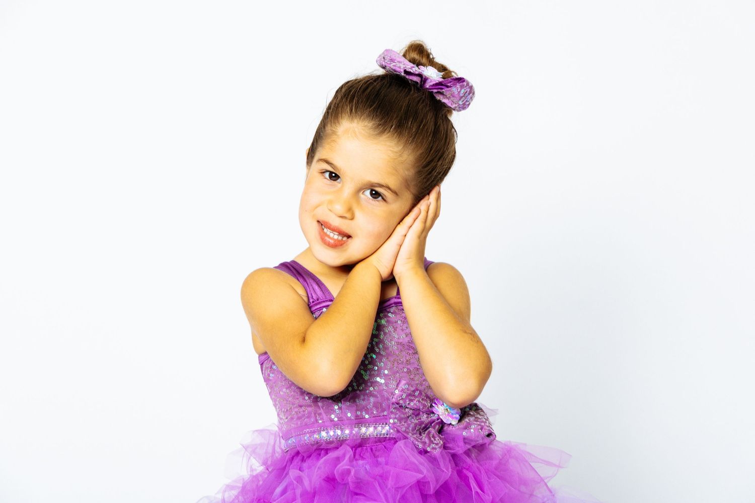 Young girl in purple tutu dress with bow poses with hands on cheeks against white background.