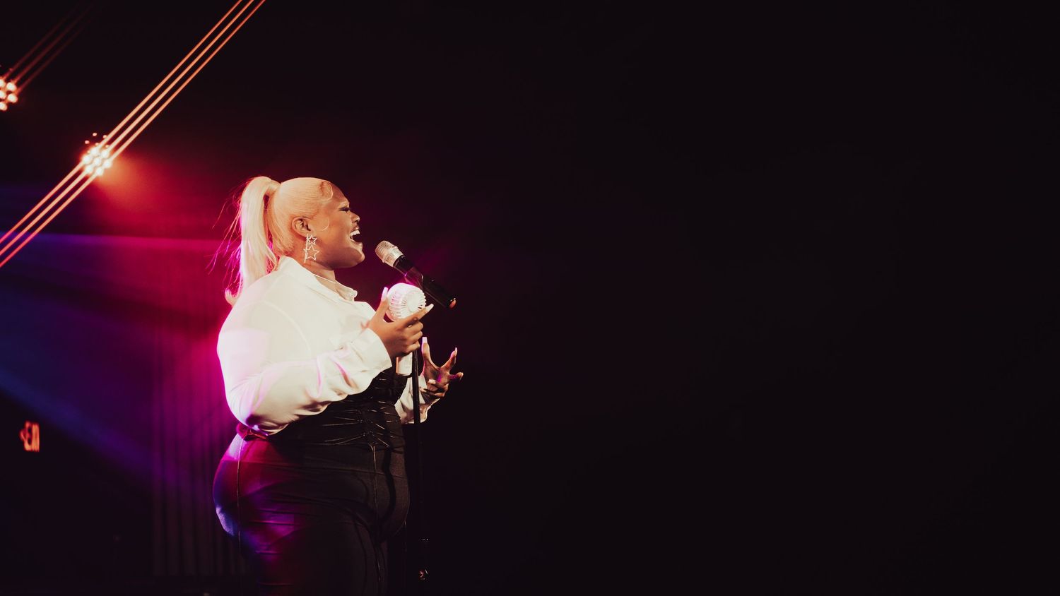 Performer with blonde ponytail sings into microphone on dark stage with dramatic red and purple stage lighting.