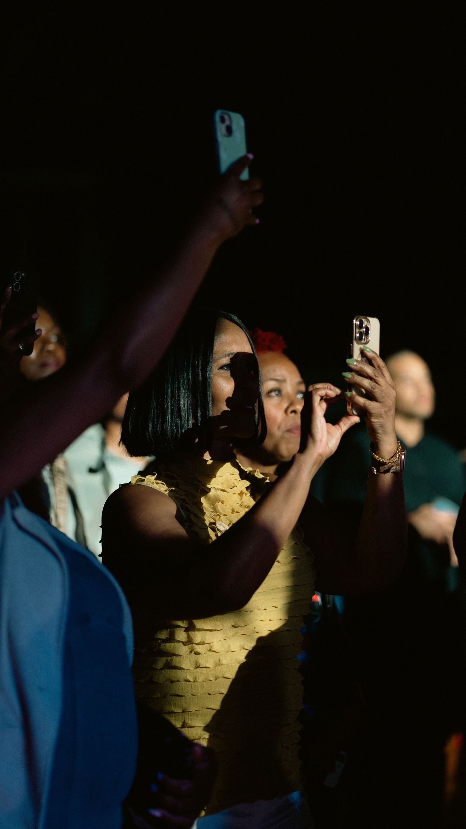 Audience members recording a live concert performance on smartphones in a dark venue with warm yellow lighting.