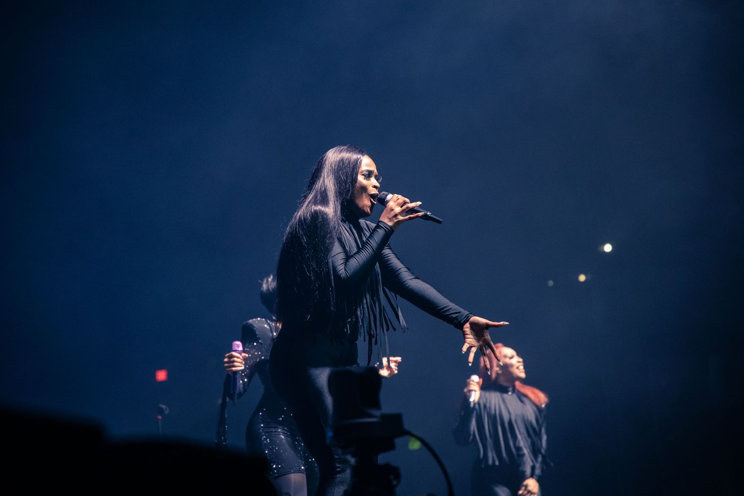 Female singer performing on stage in black outfit with backup dancers under dramatic blue stage lighting.