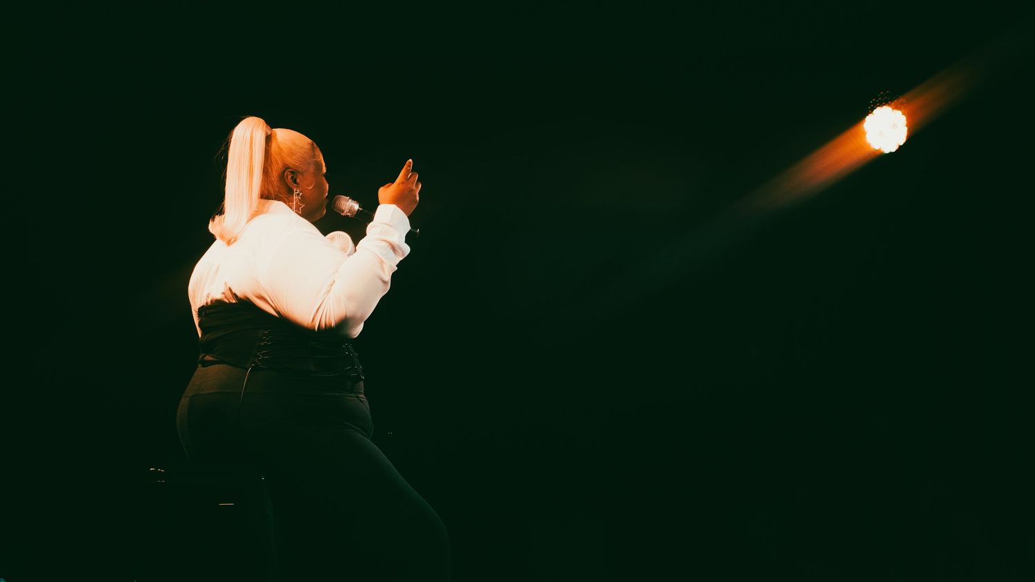 A performer in a white shirt gestures dramatically on a dark stage illuminated by a single spotlight.