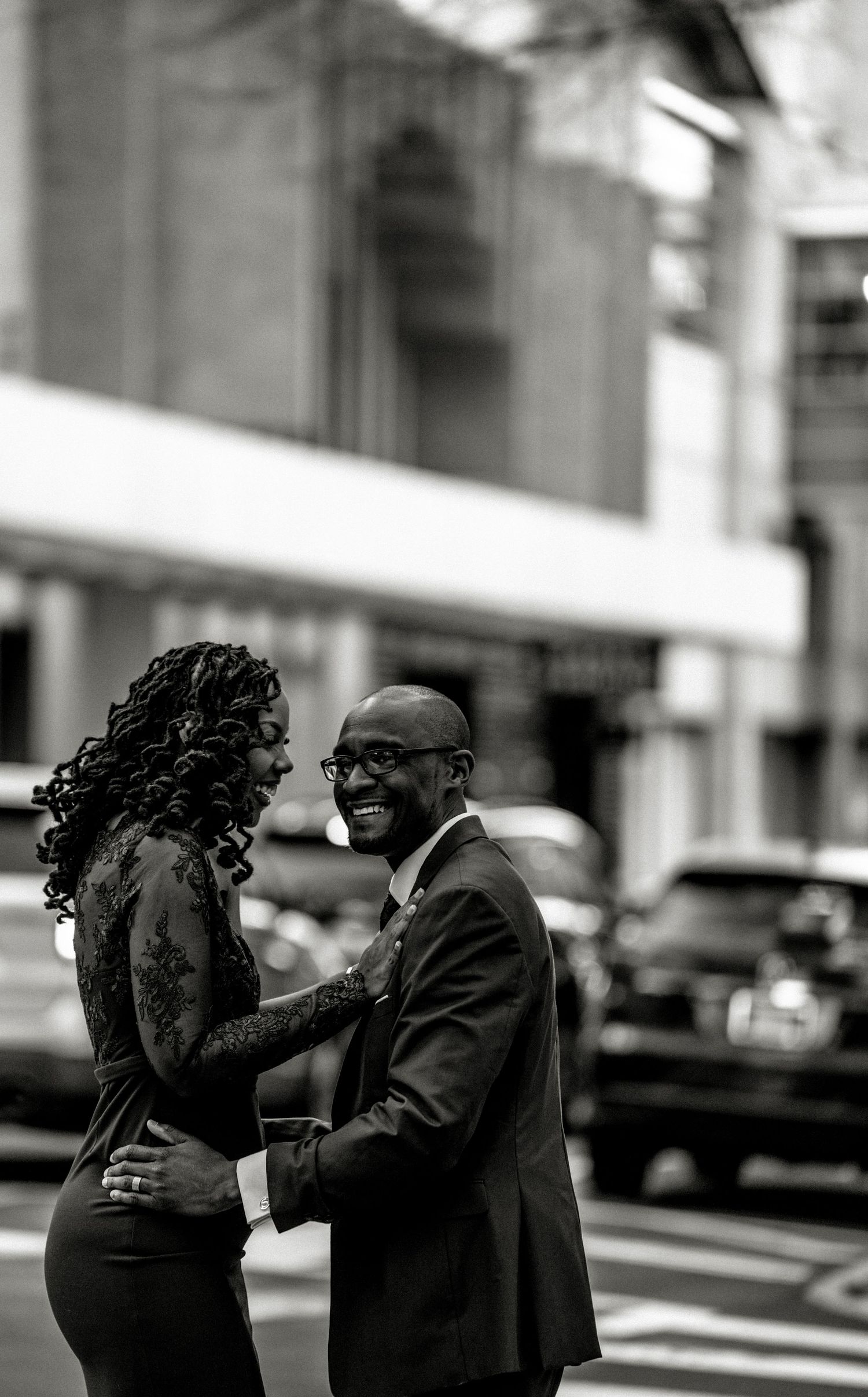 Black and white photo of a smiling couple embracing on a city street, dressed in elegant formal attire.