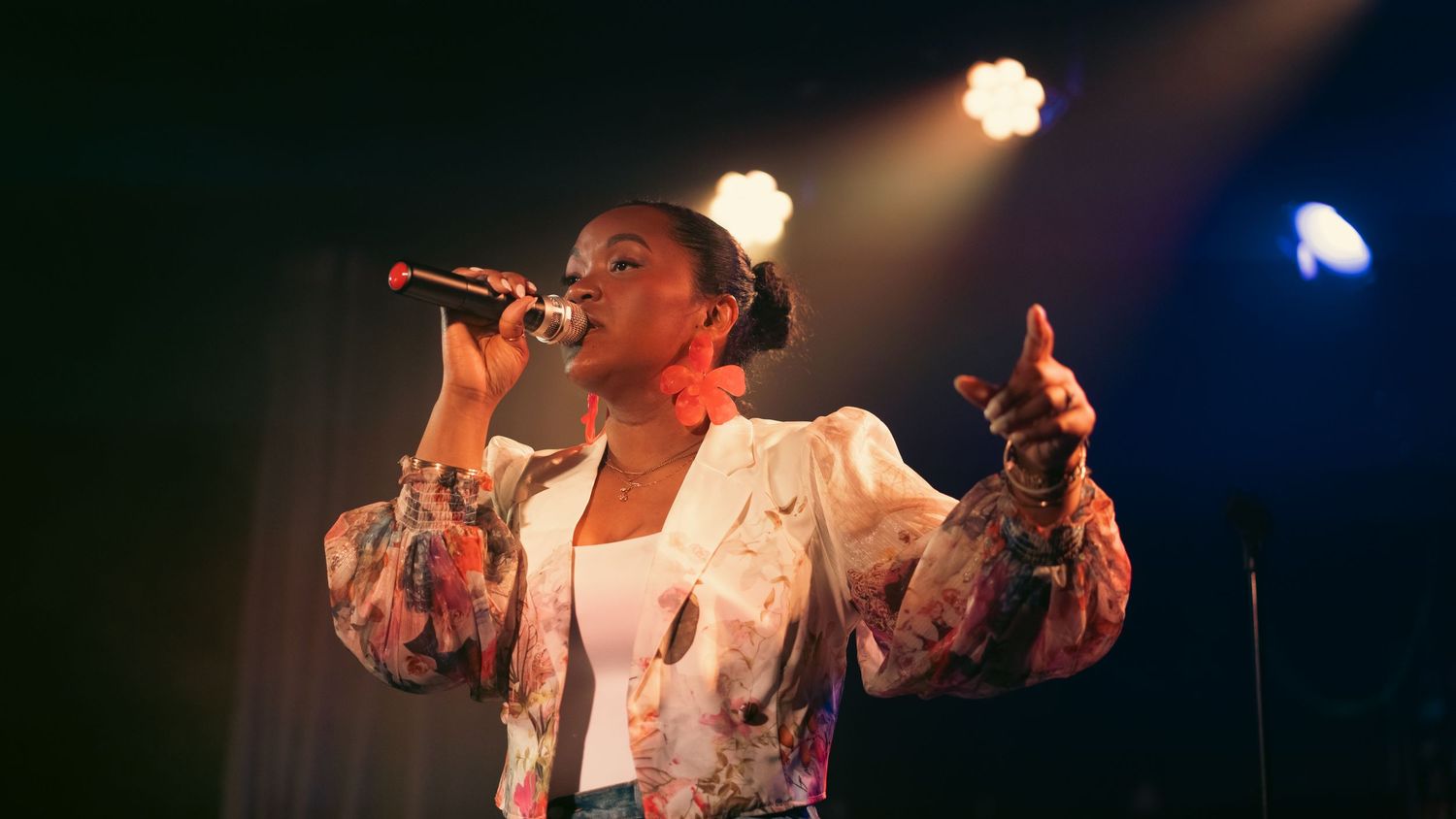 Female singer performing on stage, holding microphone, wearing floral jacket under bright stage lights.