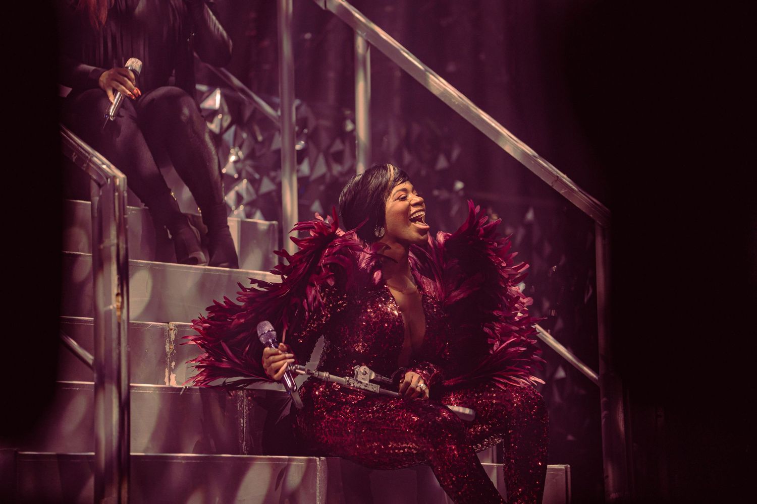 A performer in a stunning red feathered costume sings passionately on a glittering staircase stage set.