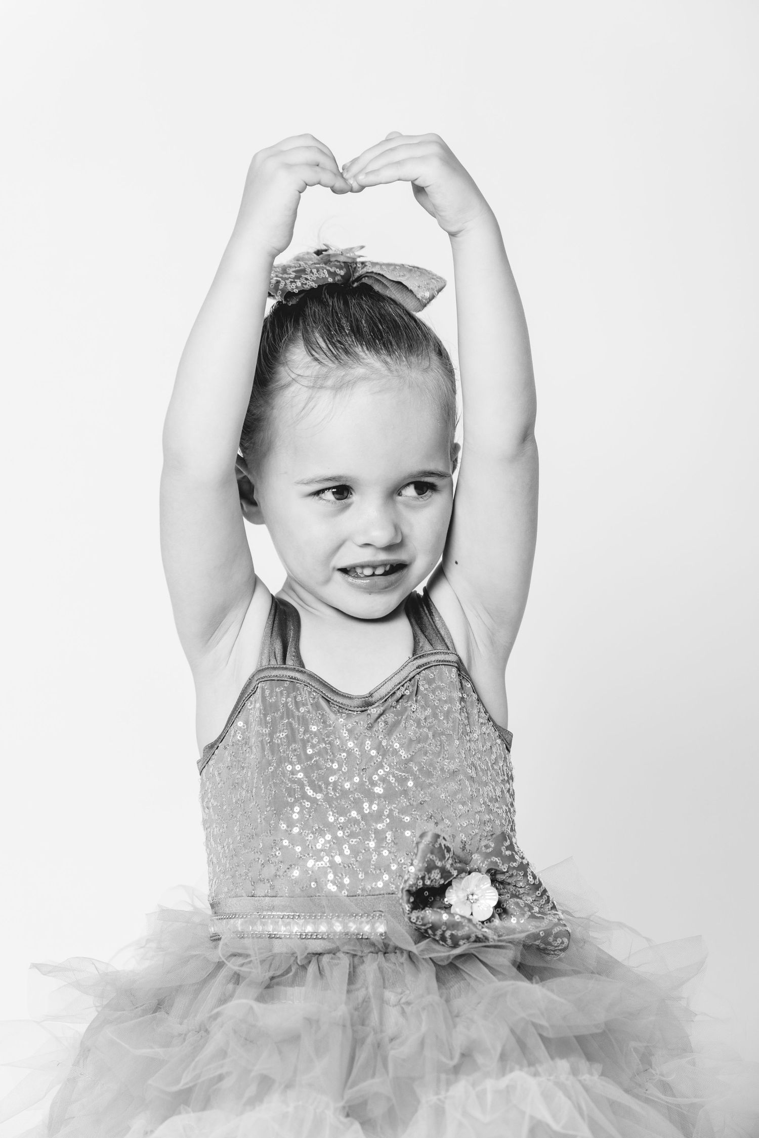 Young girl in sequin ballet costume makes heart shape with hands raised above head, black and white photo.