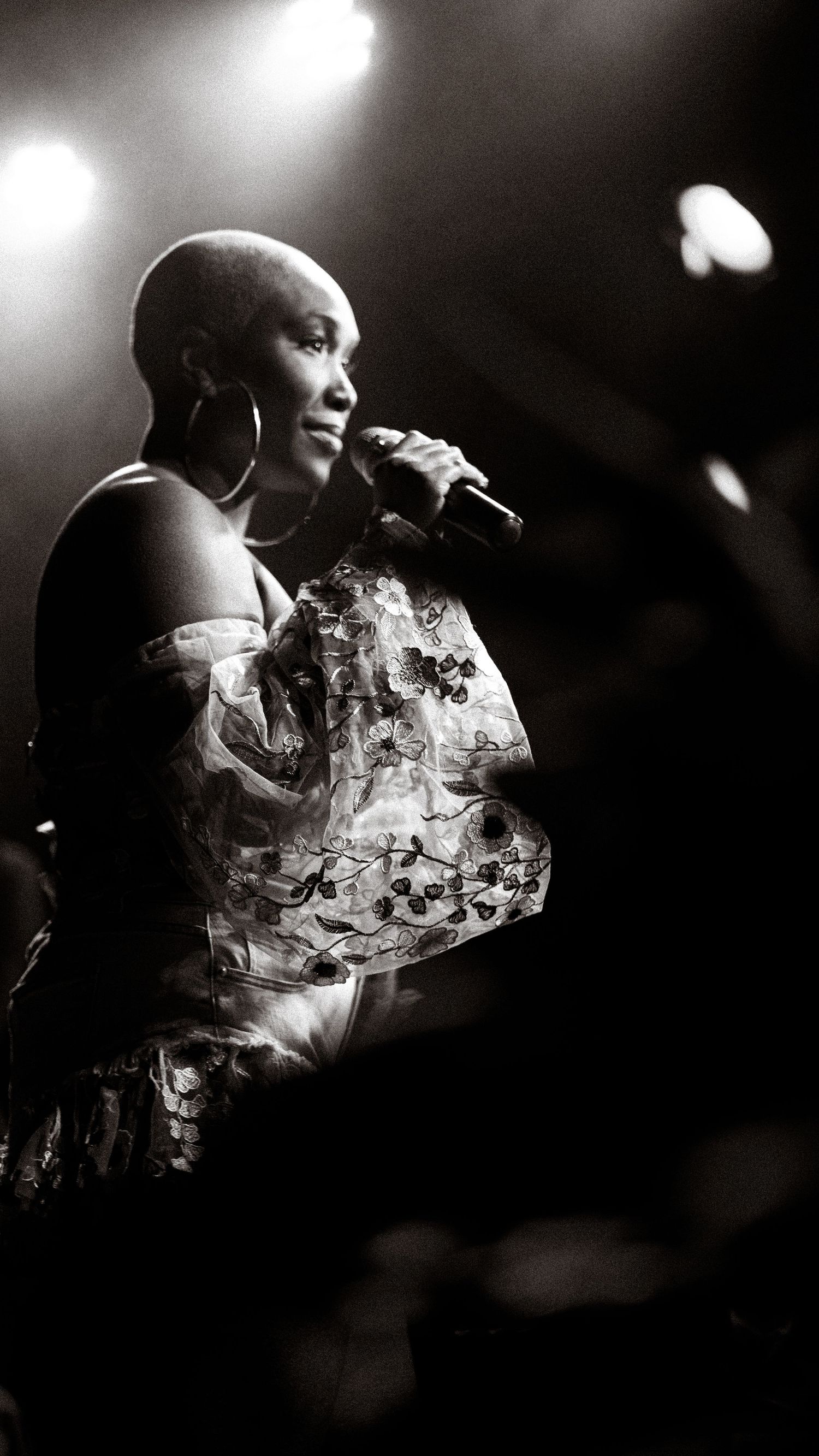 Black and white photo of a bald woman singing into a microphone on stage wearing a floral off-shoulder dress.