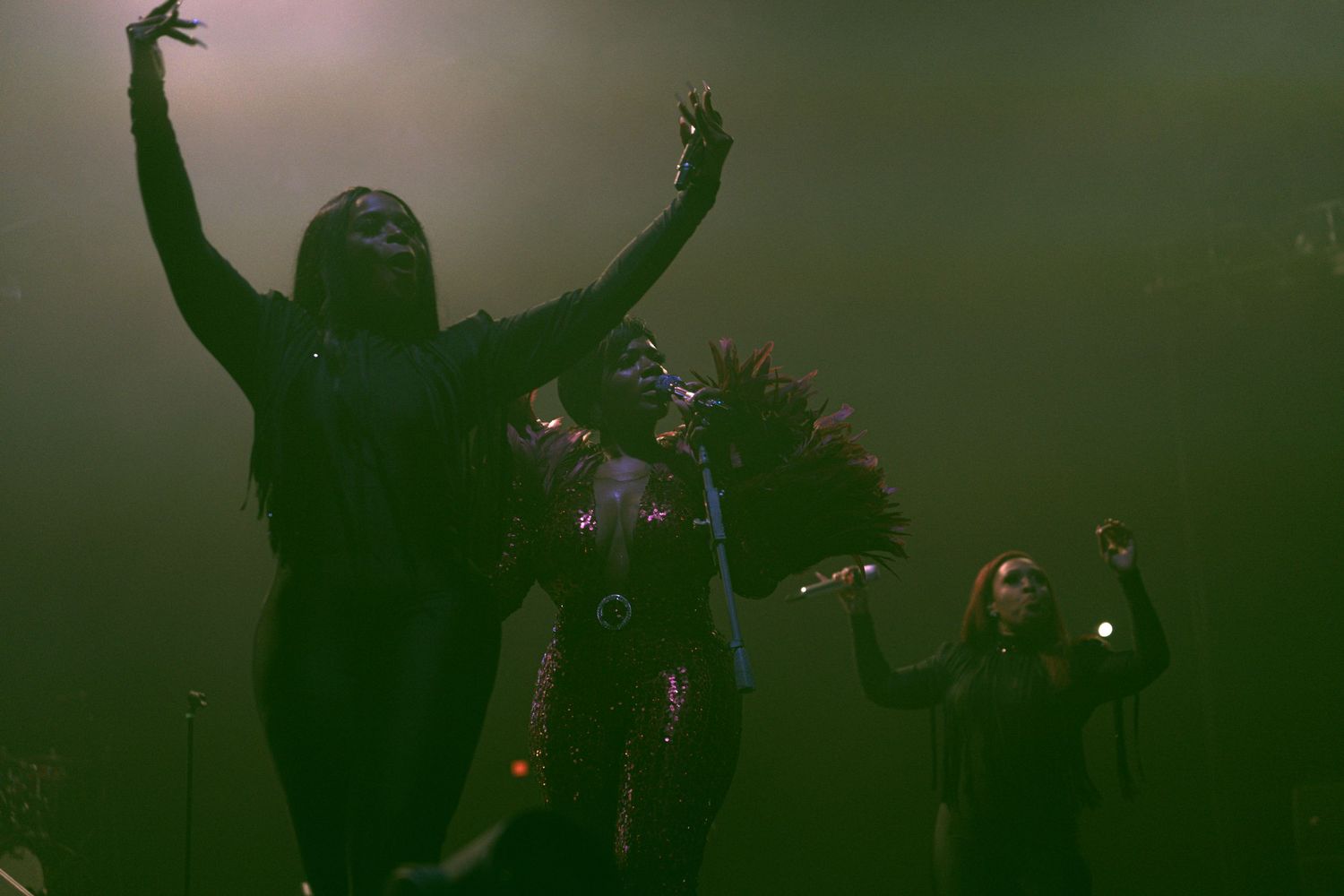 Three performers on a smoky stage with dramatic green lighting during a live concert performance.