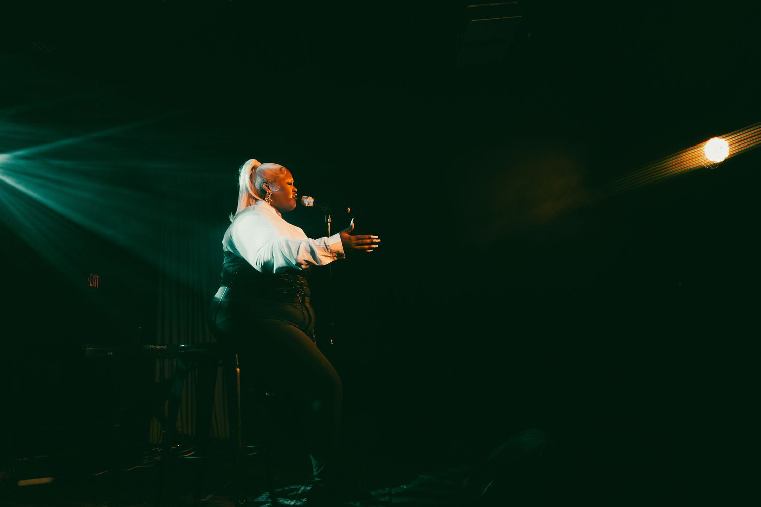 Performer on dark stage with dramatic green spotlights, wearing white shirt, arms outstretched during live concert.