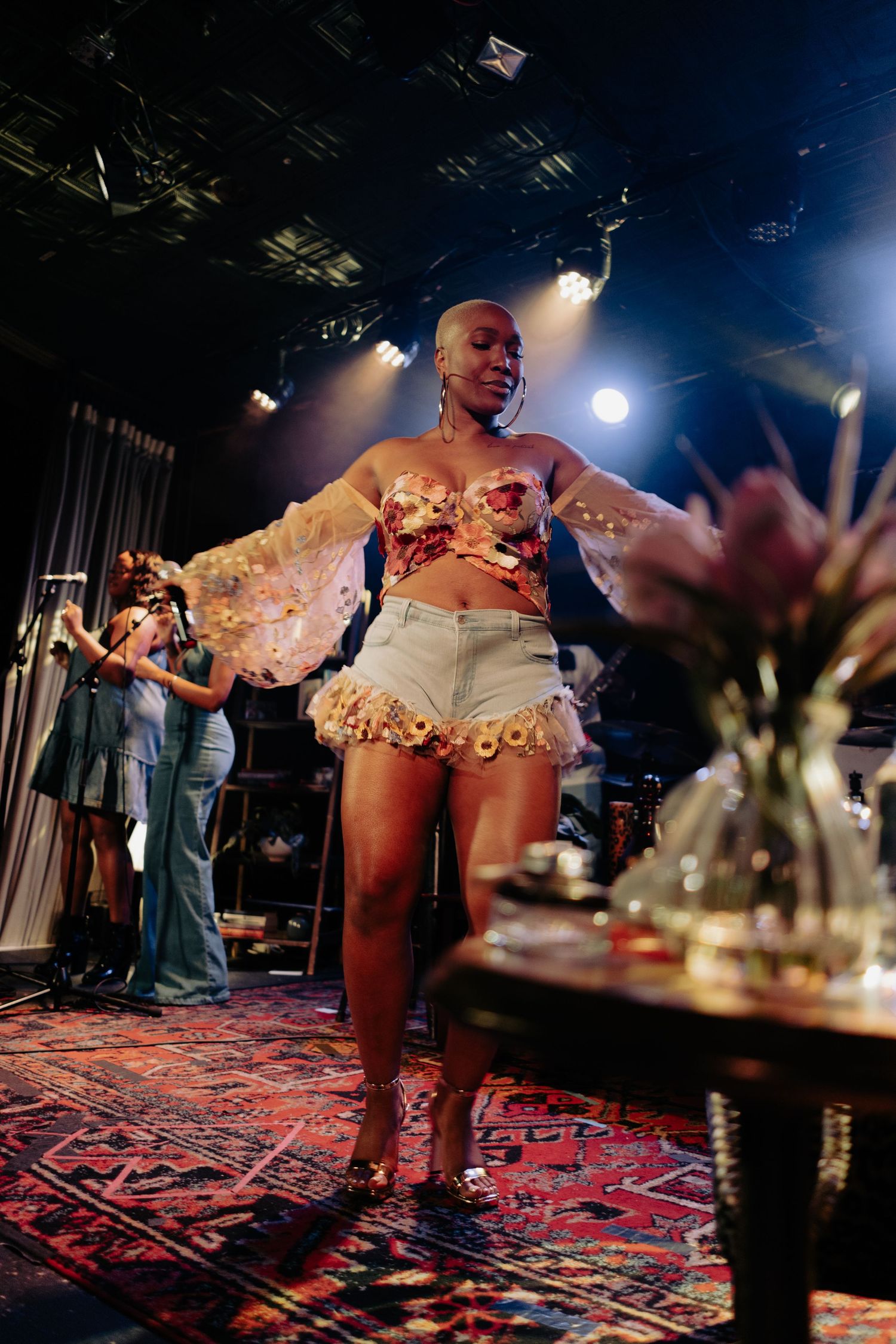 Bald female performer dances on stage in floral corset and white shorts under spotlights.