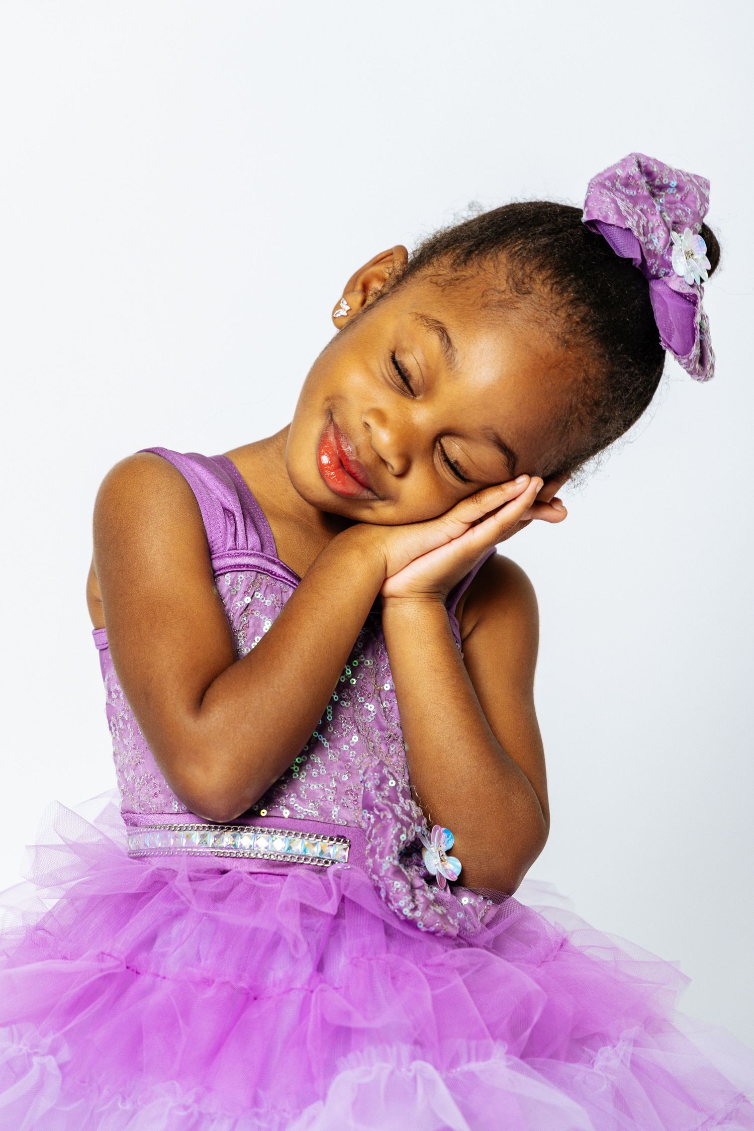 Young Black girl in purple sequin tutu dress with bow, eyes closed, hands on cheeks, smiling peacefully.