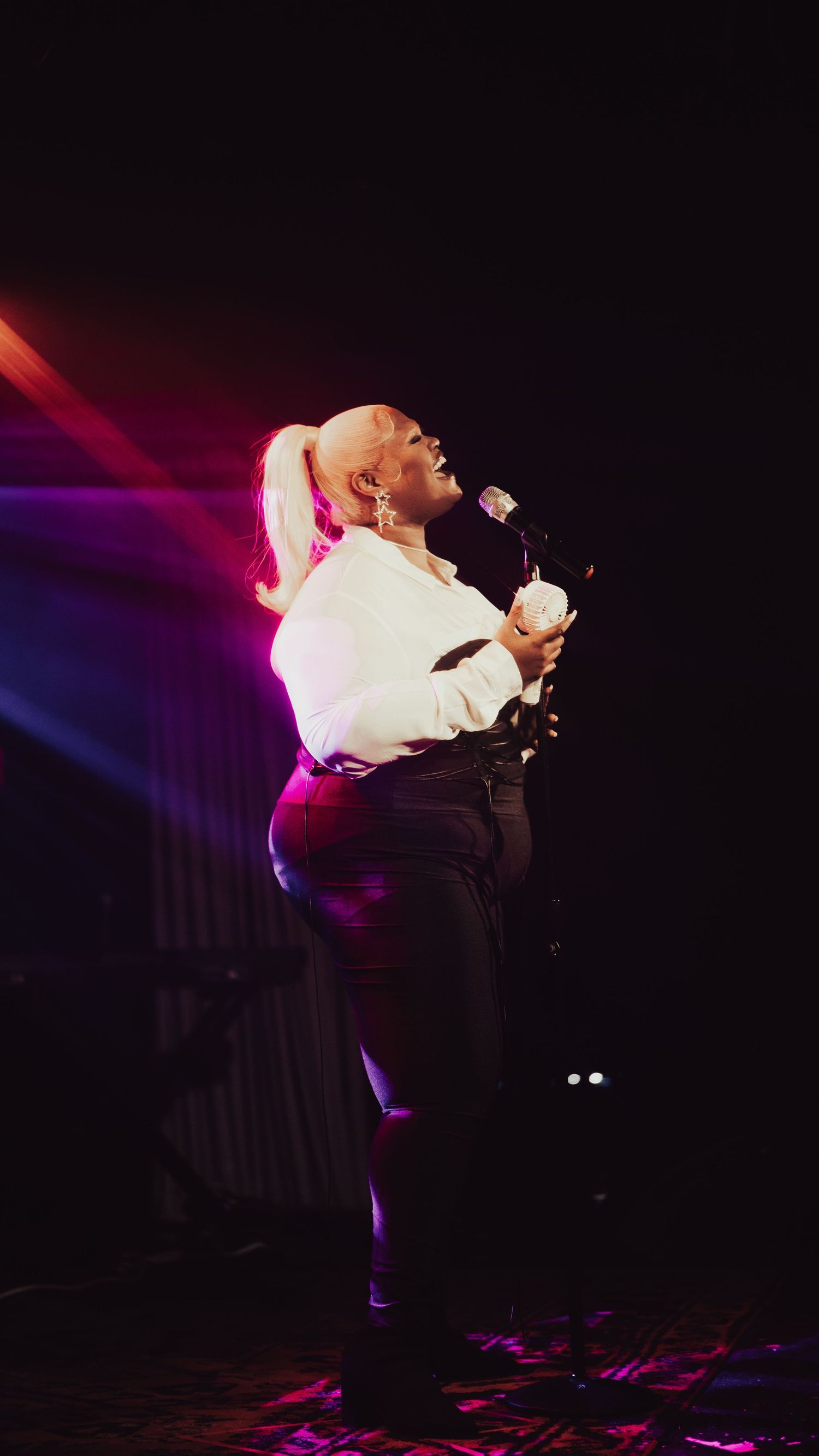 A female singer performs on stage in a white blouse and black pants, holding a microphone under dramatic pink stage lighting.