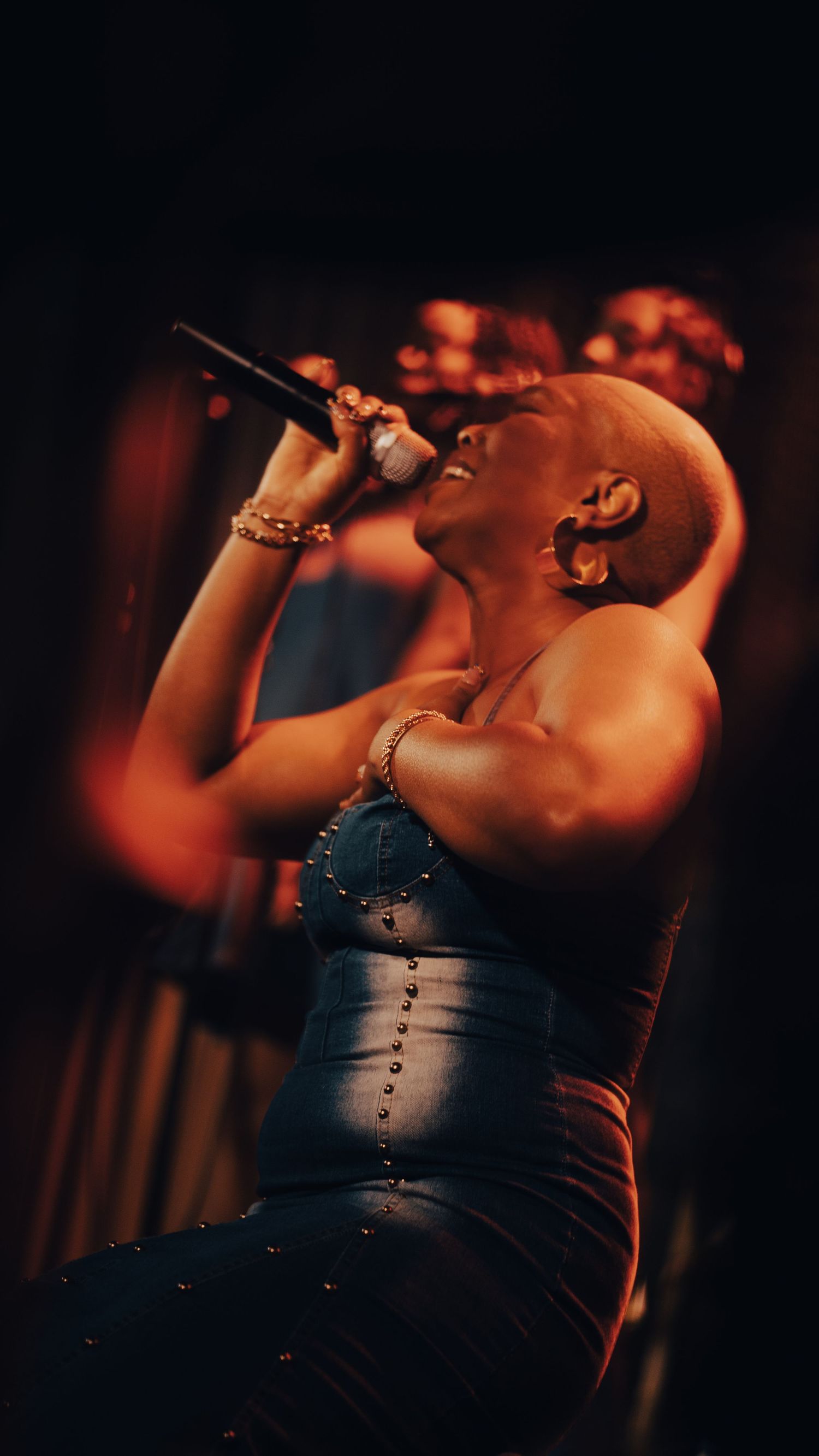 Bald female vocalist singing passionately into microphone wearing blue studded corset under warm orange stage lighting.
