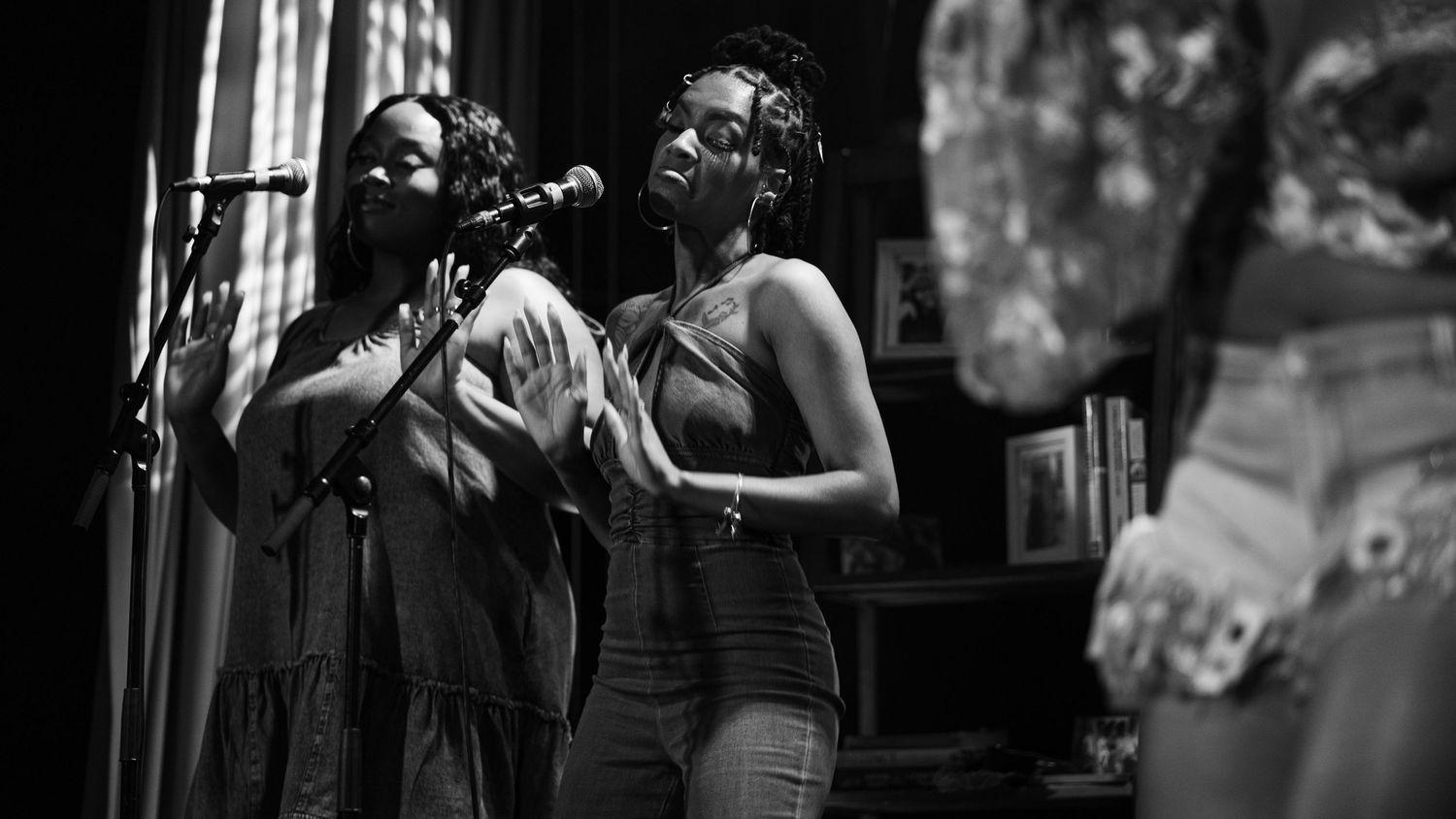 Two female vocalists sing together at a microphone in a black and white concert photo.