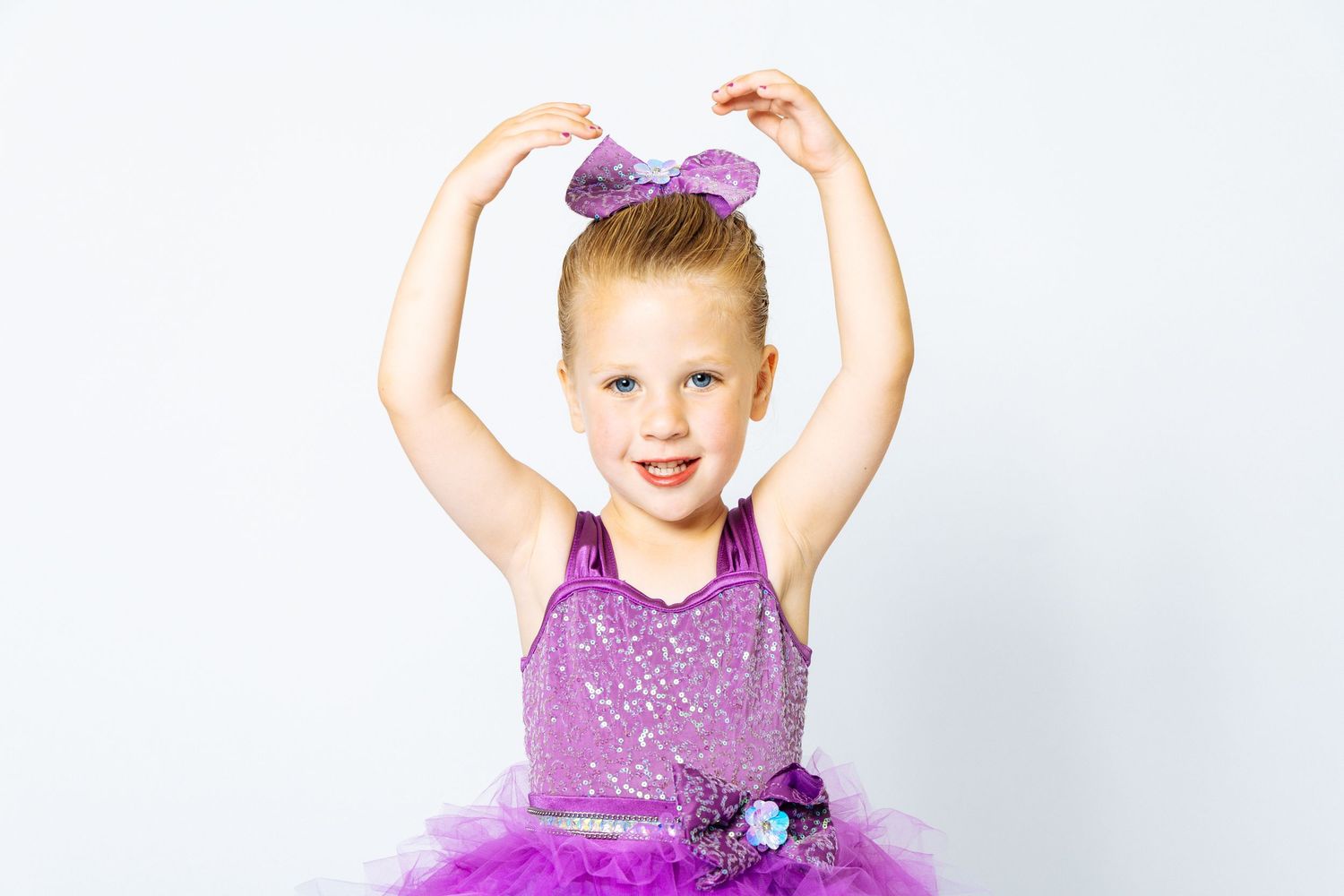 A young girl in a purple sequin tutu dress with a bow headband poses with arms raised on white background.