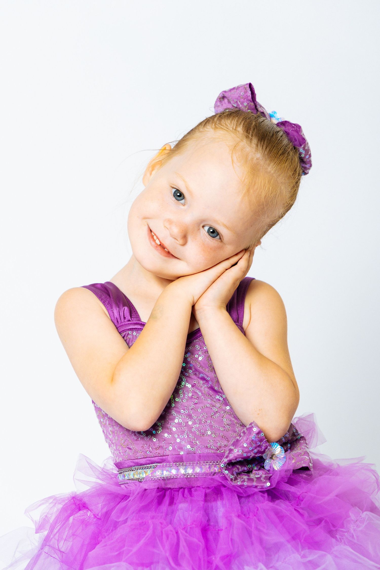 Smiling blonde toddler girl in purple sequin tutu dress with bow, hands on cheeks, blue eyes, white background.