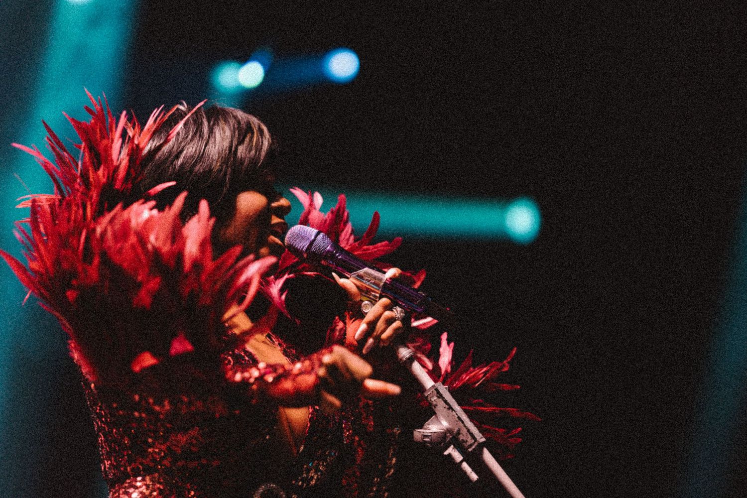 Singer performing on stage wearing a dramatic red feathered costume, holding a microphone under blue stage lights.