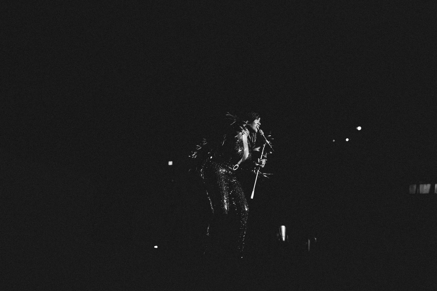 Black and white concert photo of a performer on stage with microphone stand under dramatic spotlight at night.