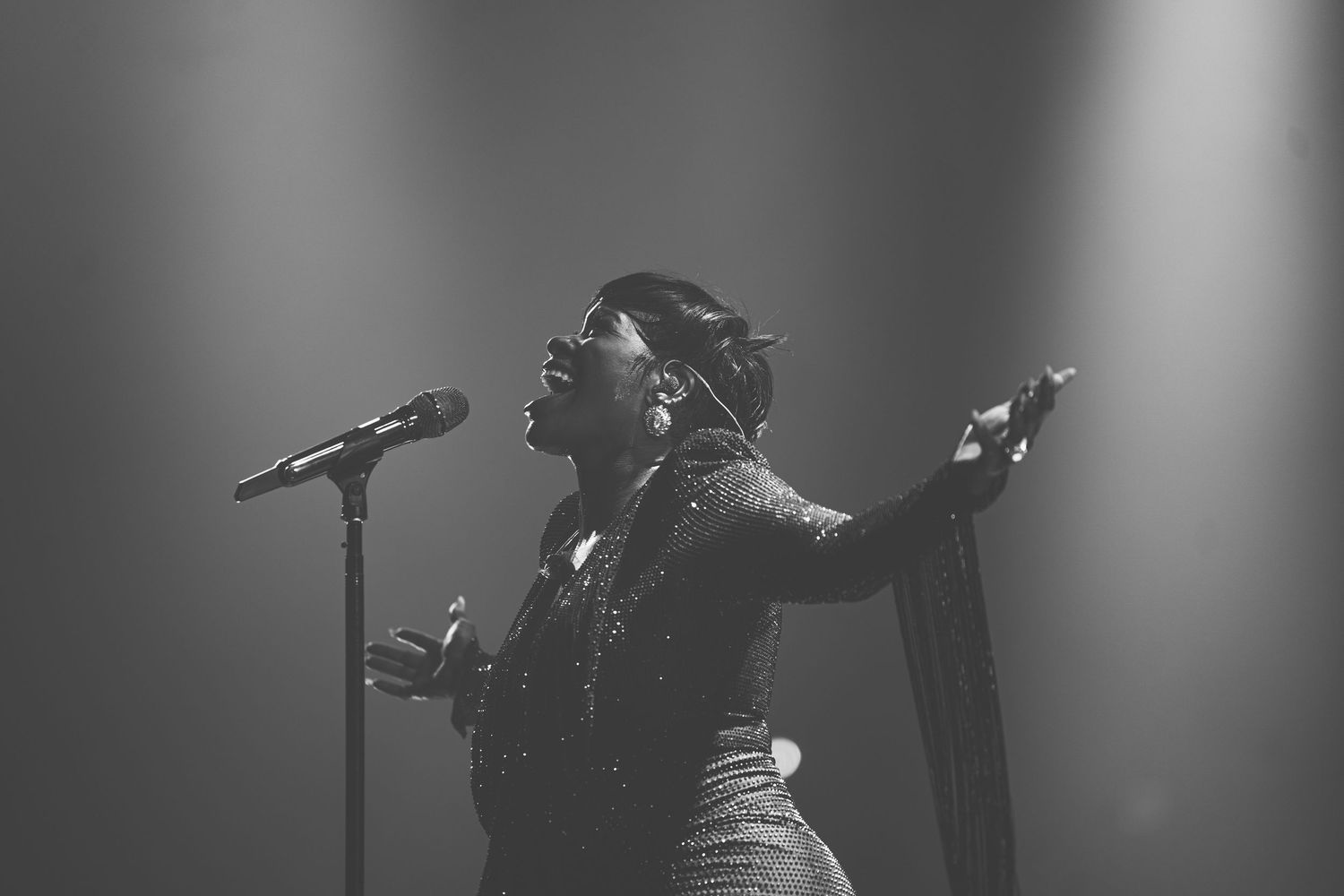 A powerful vocalist performs on stage in a sequined outfit with arms outstretched under dramatic spotlights.