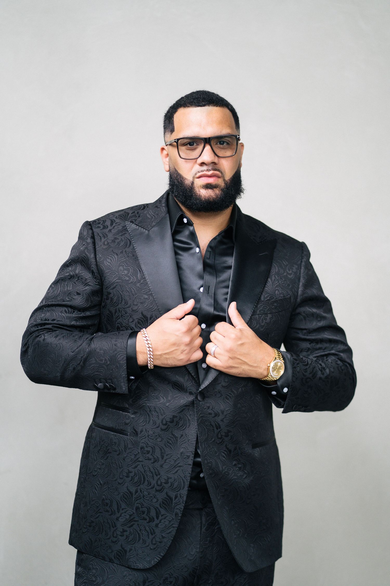 Professional studio portrait of person in black textured suit jacket and glasses.