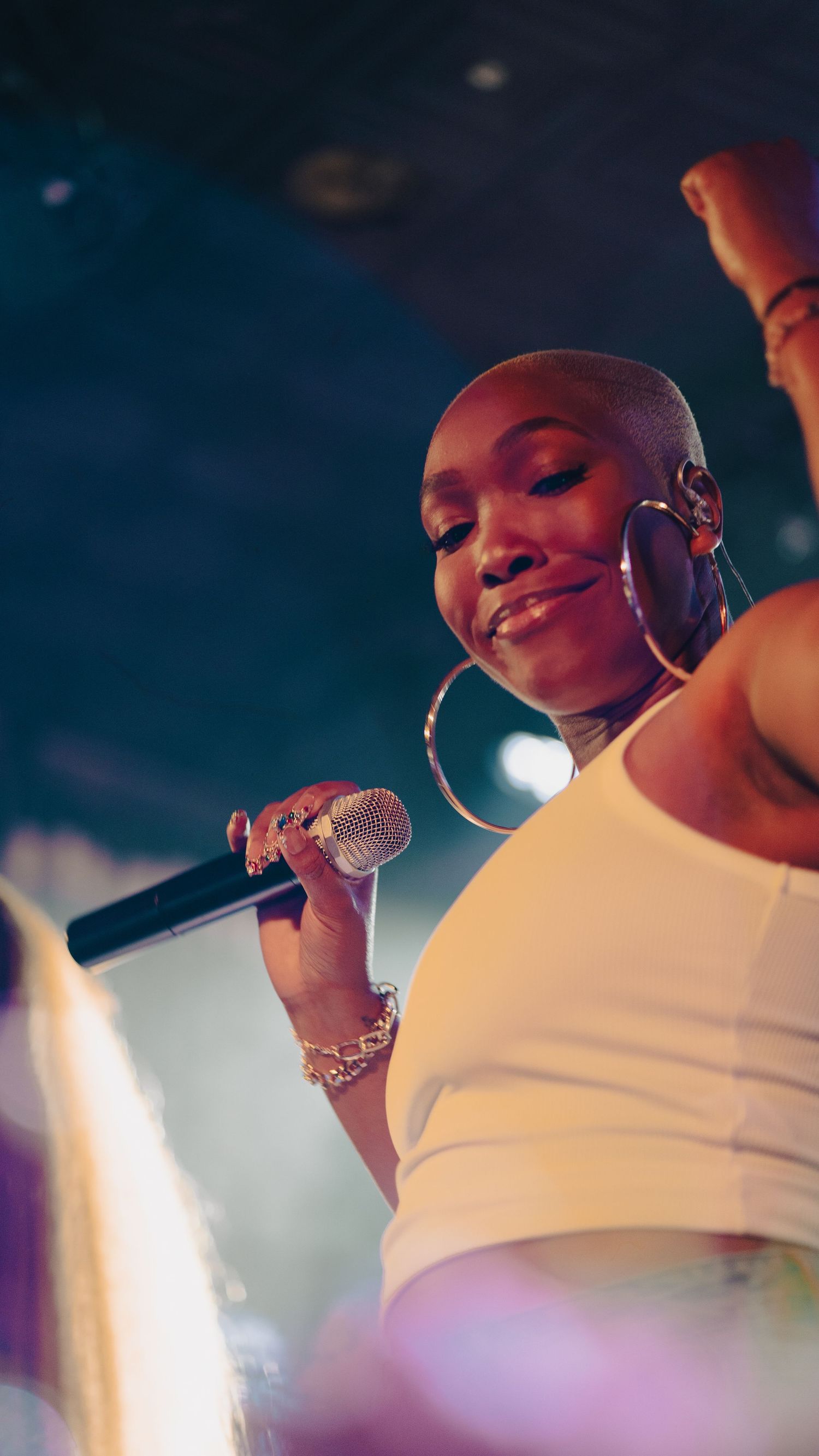 Bald female singer performing on stage holding microphone wearing white top and large hoop earrings under blue stage lights.
