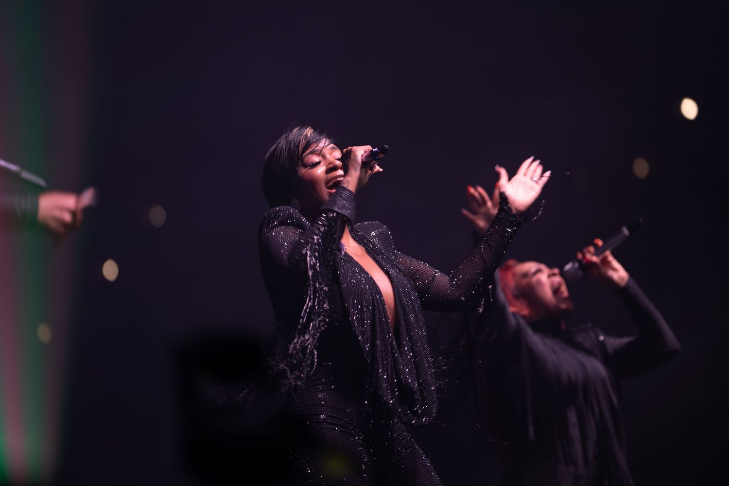 A female singer in a black sequined outfit performs passionately on stage with backup singers.