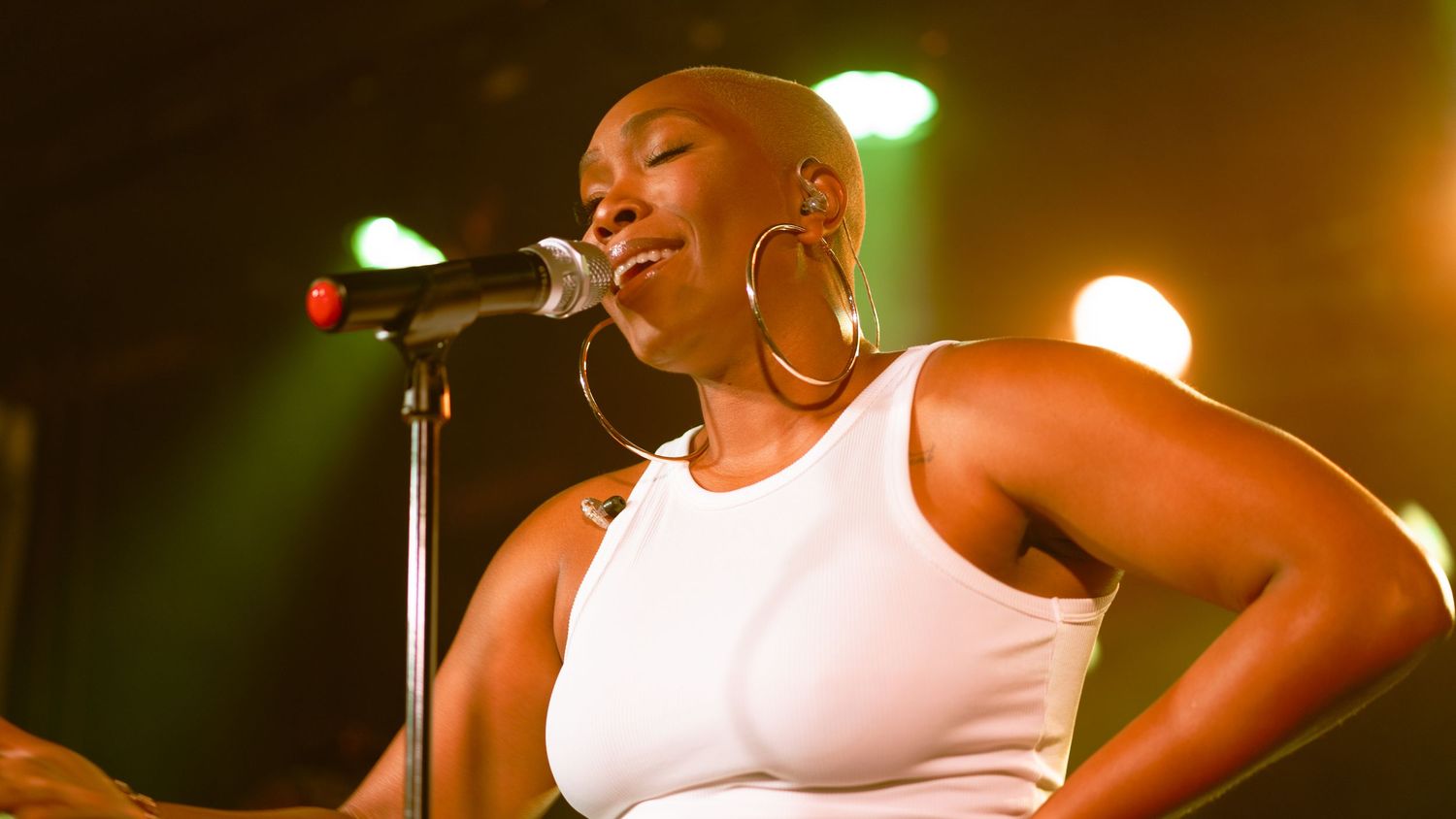 Bald female vocalist singing on stage wearing white top with large hoop earrings under warm stage lights.