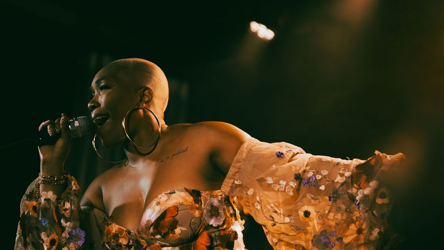 A bald female singer performs on stage, wearing a floral gown and large hoop earrings, singing into a microphone under dramatic lighting.