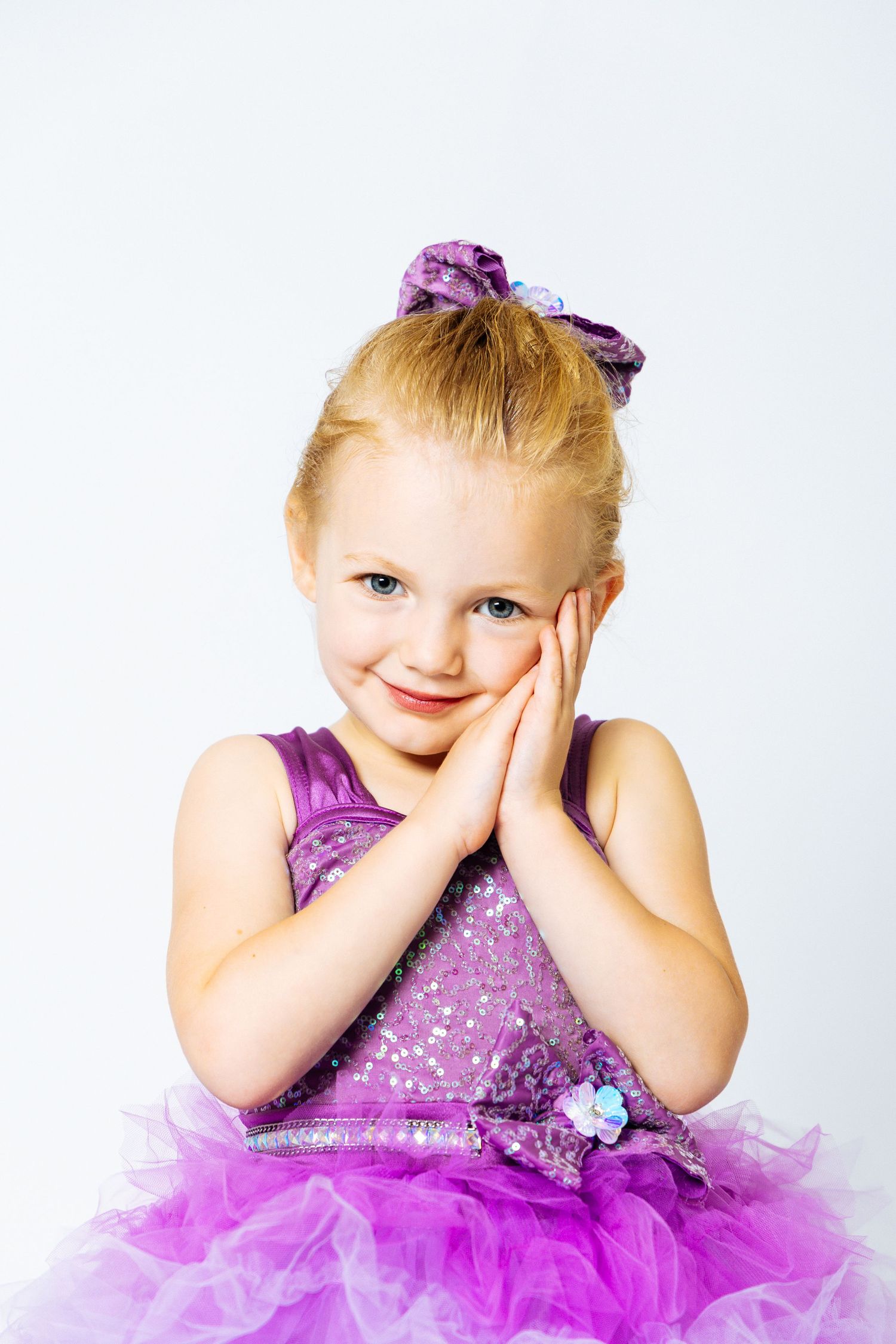 Adorable little girl in purple sequin tutu dress with matching bow poses with hands on cheeks, smiling.