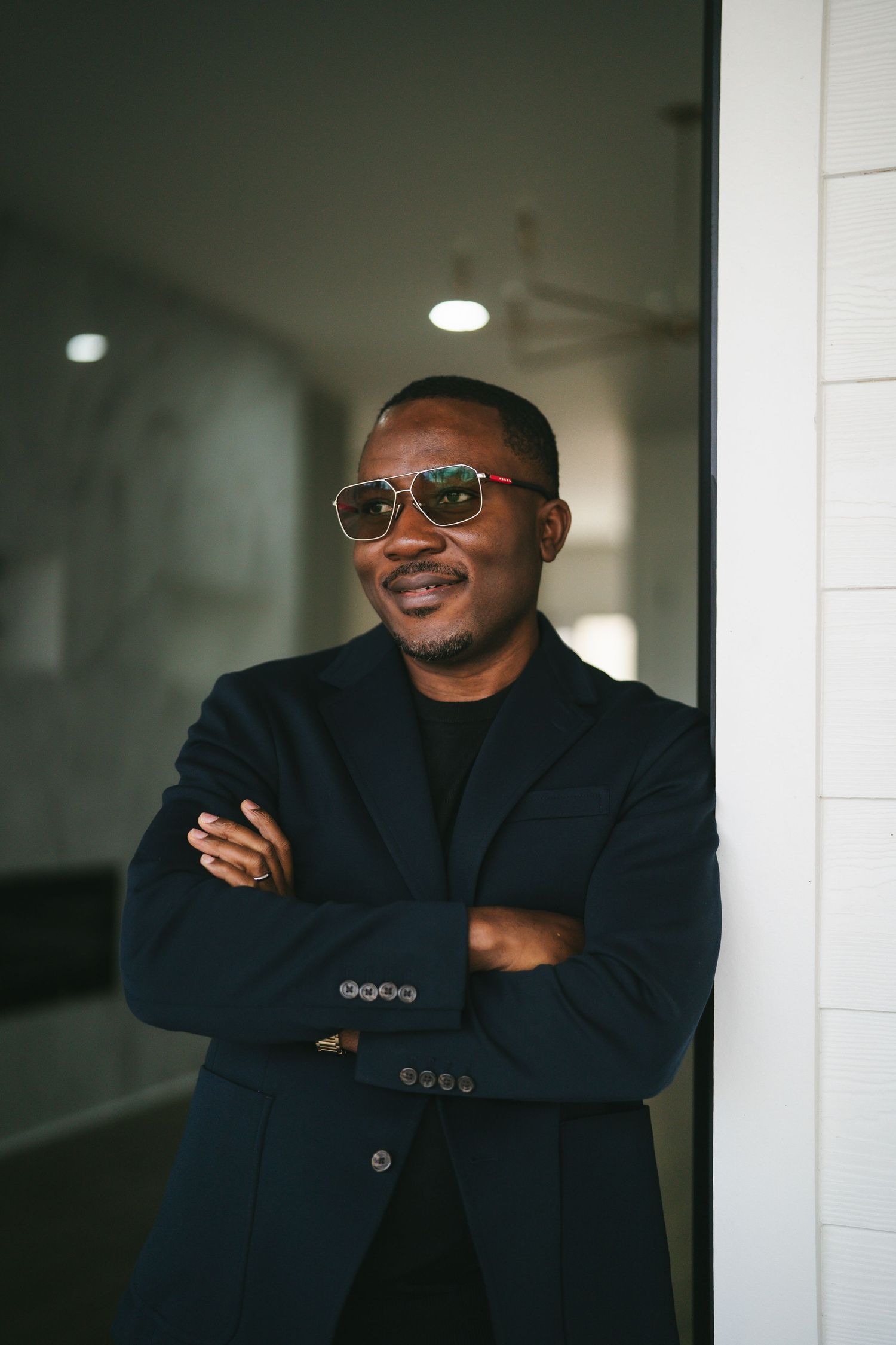 A person wearing a black blazer and sunglasses smiles while leaning against a white wall in a hallway.