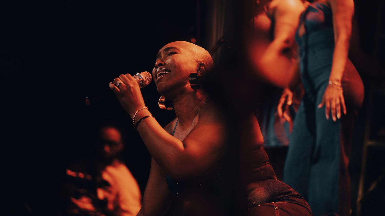 Female vocalist singing passionately into microphone with backup dancers on a dimly lit stage.