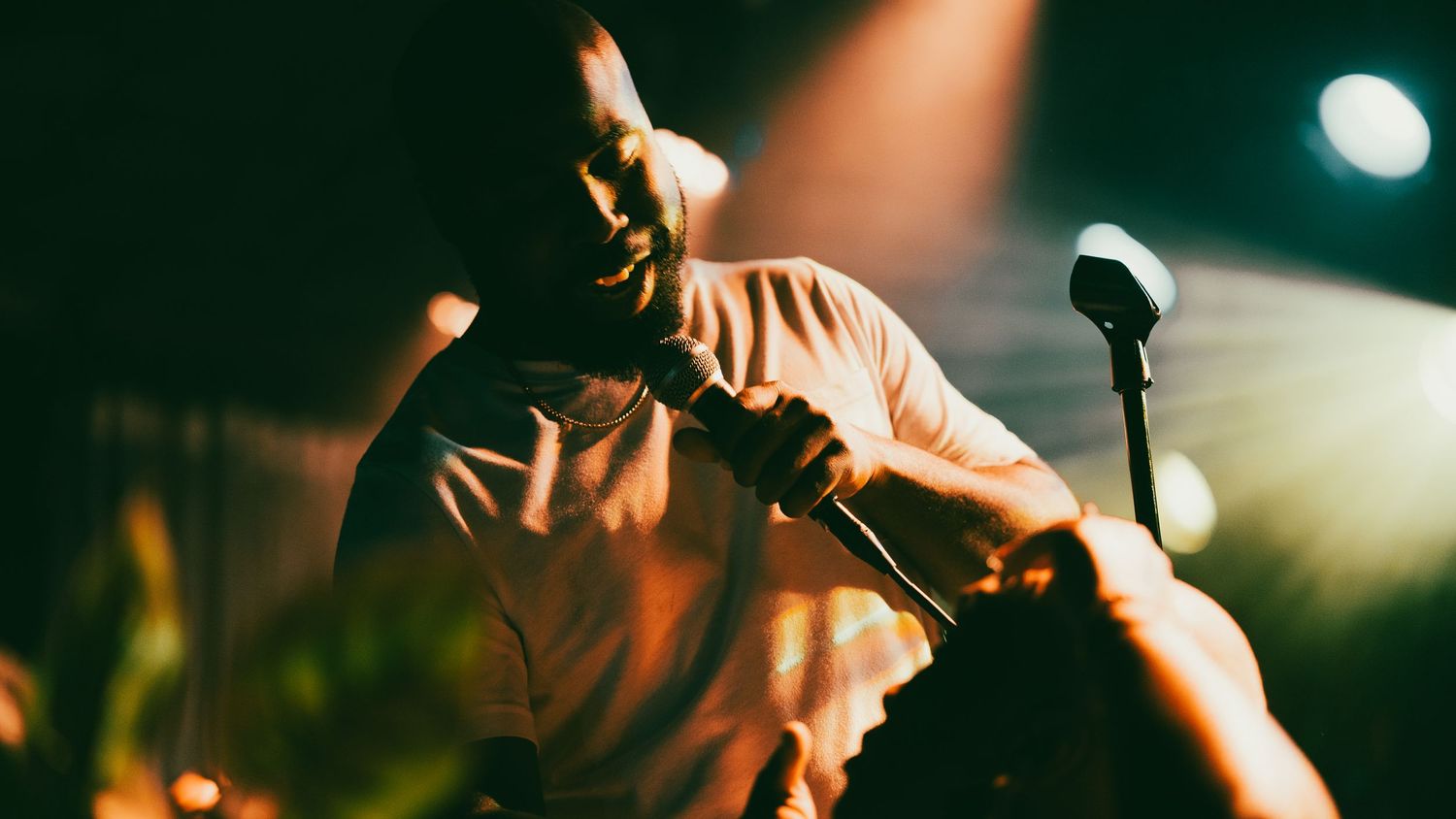 Male performer leans forward gripping microphone stand on dimly lit concert stage under warm spotlight.