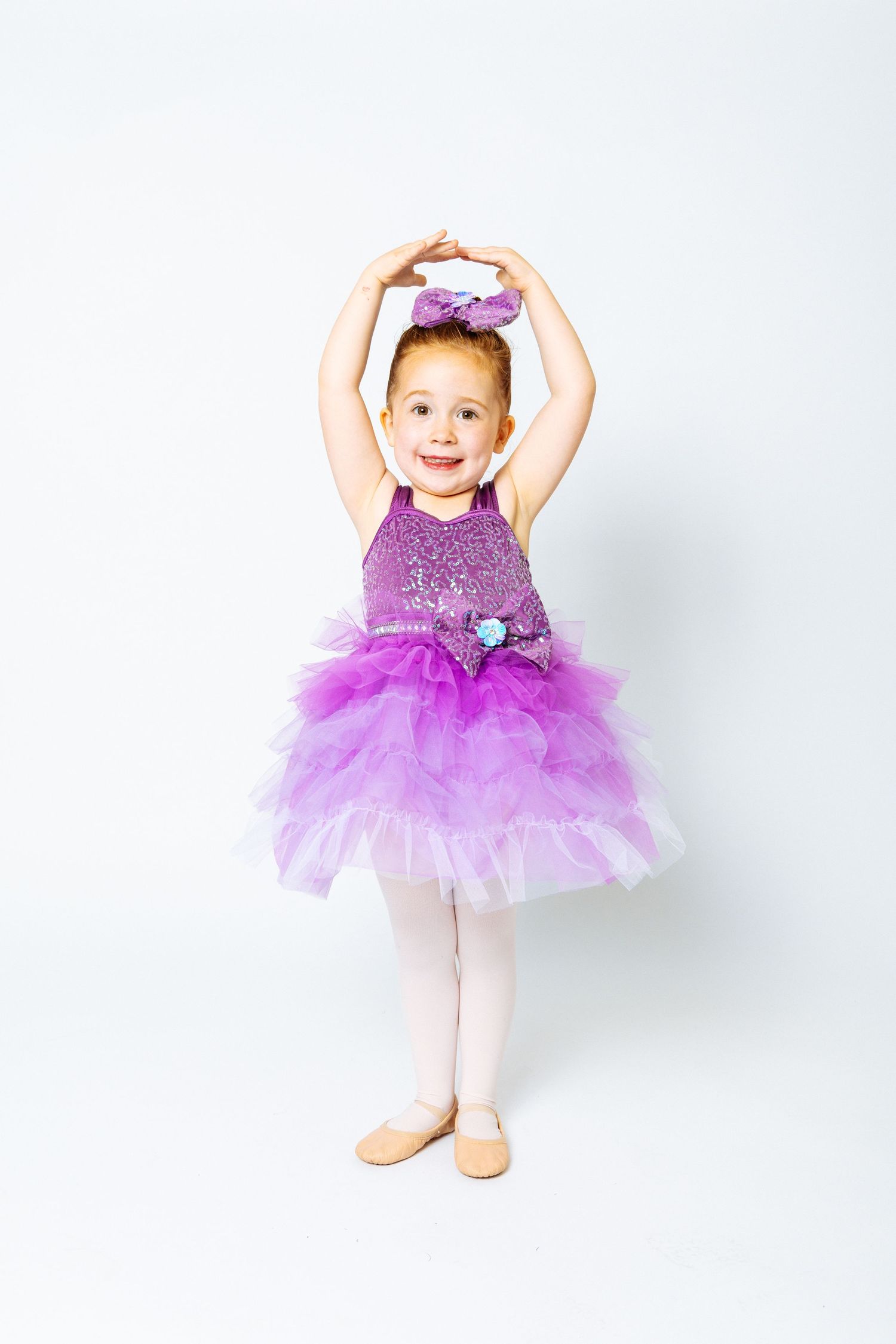 A young girl in a purple sequin ballet tutu dress strikes a dance pose with arms raised overhead on white background.