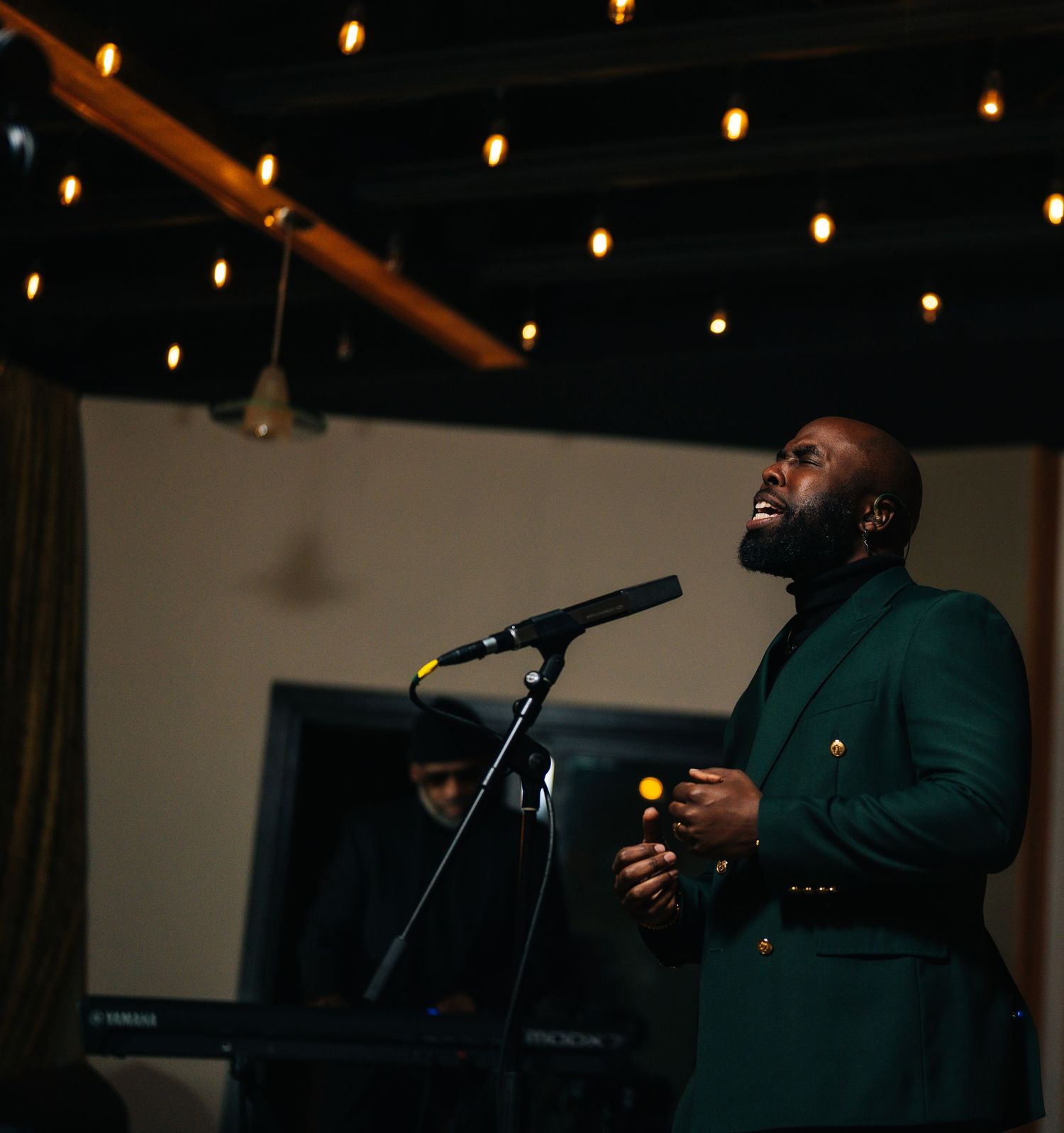 A man in a green blazer sings passionately at a microphone under warm string lights indoors.